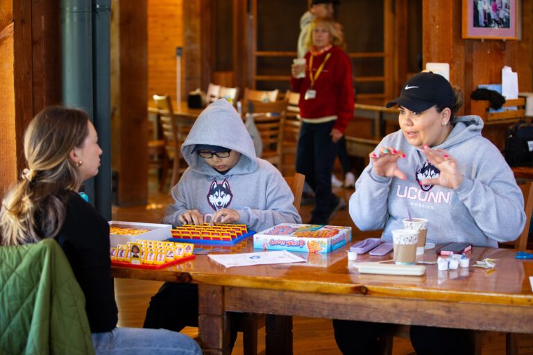 Three people sit at a wooden table playing board games inside a cozy room, while another person stands in the background. The people at the table are focused and engaged in their activity.