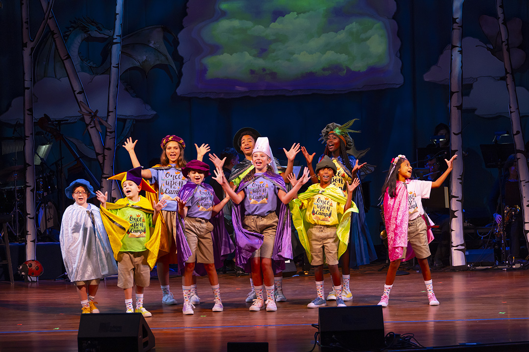 A group of children and adults perform on stage wearing colorful capes, hats, and khaki shorts. They stand in front of a whimsical backdrop with clouds and a dragon, raising their arms and smiling as part of a lively musical scene.