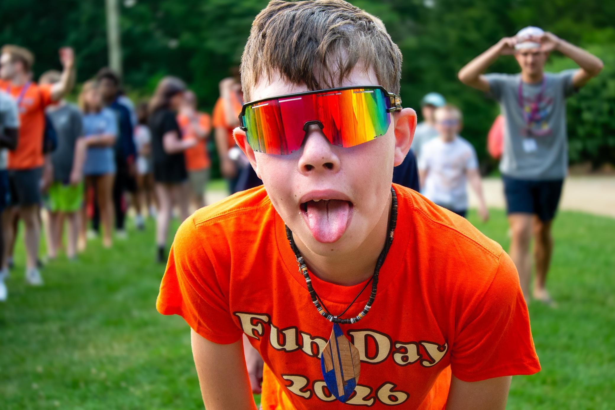 A boy in rainbow sunglasses and an orange Fun Day 2026 shirt sticks out his tongue at the camera, with a group of people blurred in the background on a grassy field.