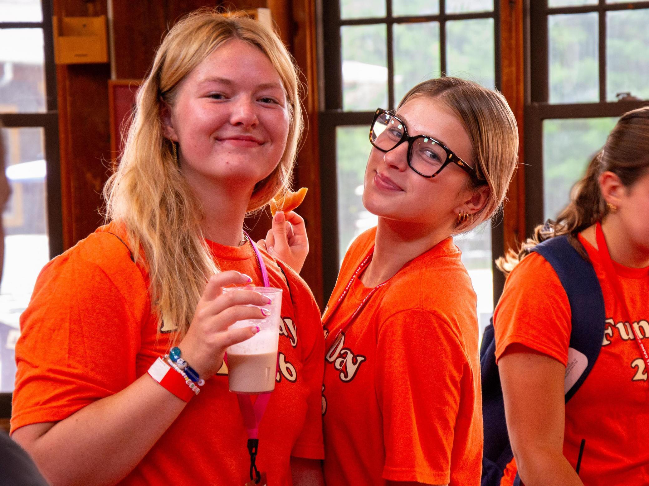 Two smiling teens in orange shirts stand indoors; one holds a drink, and the other wears black glasses. Bright sunlight shines through windows behind them.