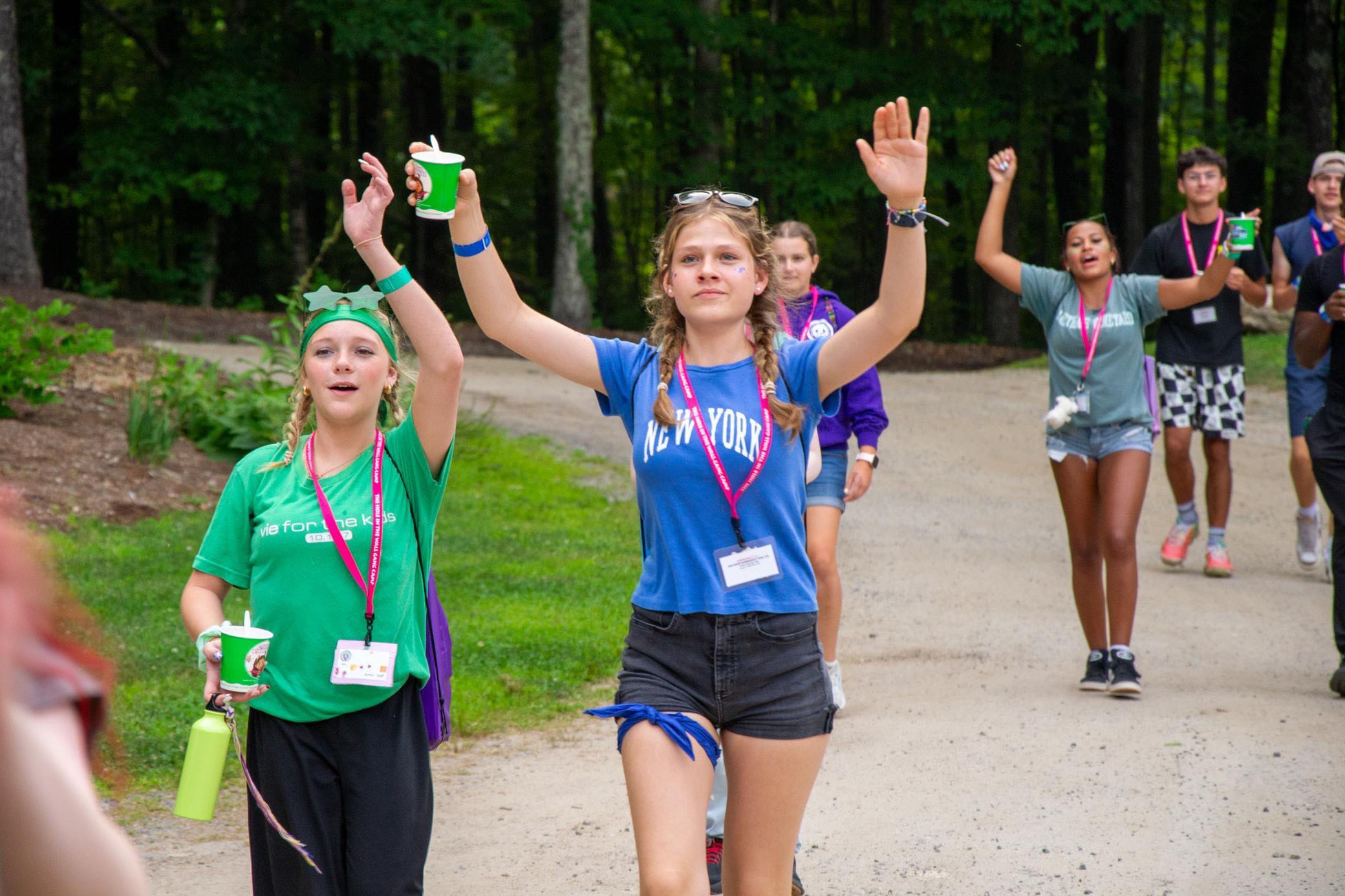 A group of teenagers walk outdoors on a dirt path, raising their arms and smiling. They wear colorful outfits and lanyards, with trees and greenery in the background.