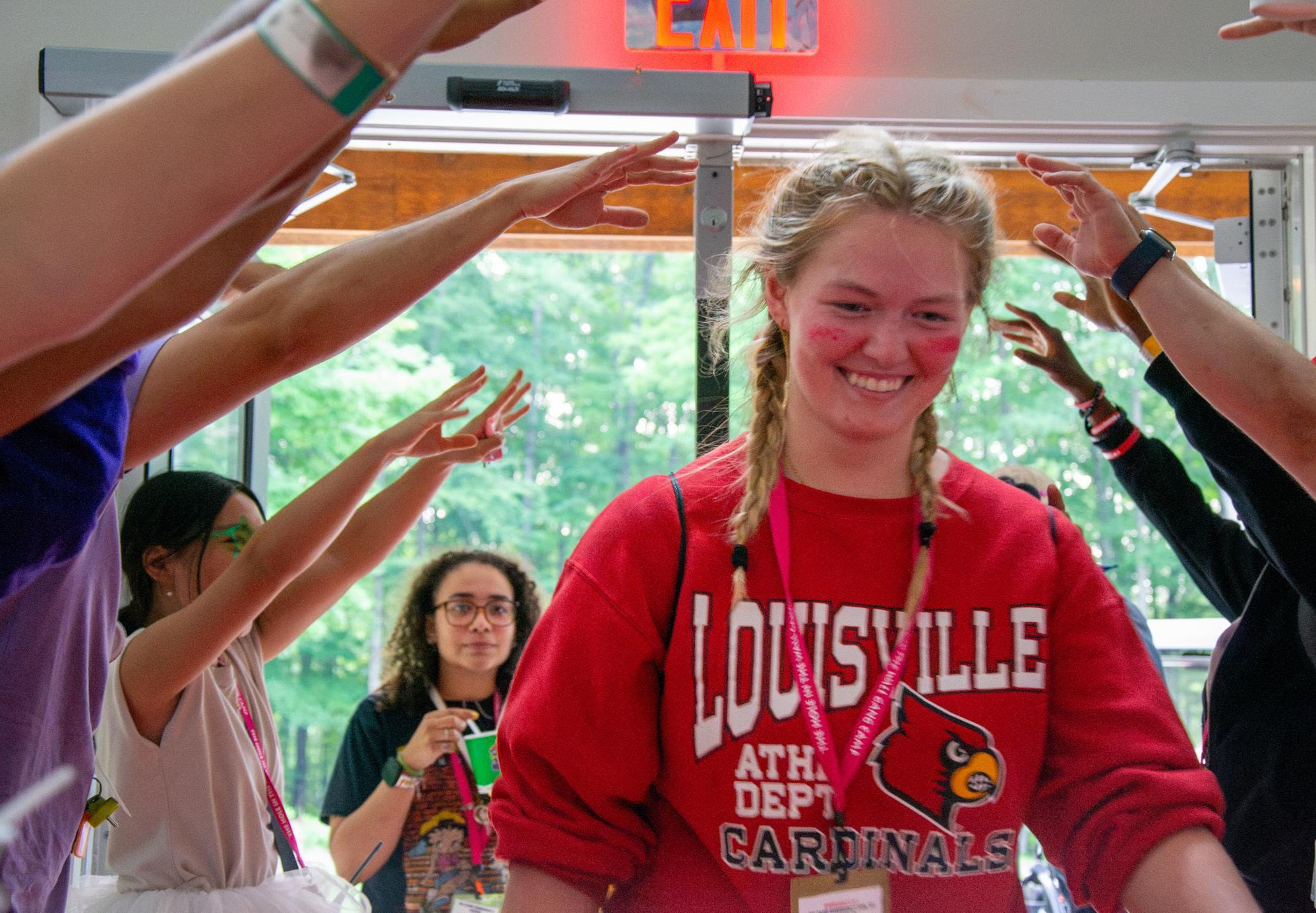 A smiling young woman in a red Louisville Cardinals sweatshirt walks through a tunnel of raised arms indoors, with two people in the background watching.