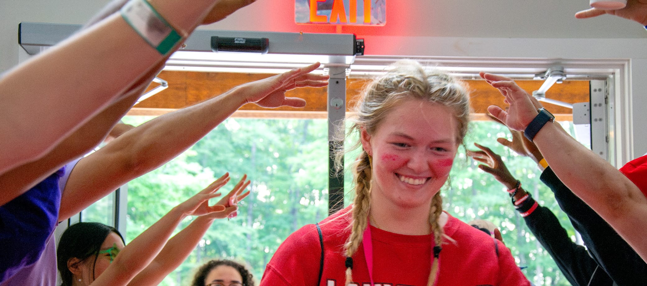A smiling young person with face paint walks indoors under an arch of raised hands, with sunlight and greenery visible through open doors behind them.