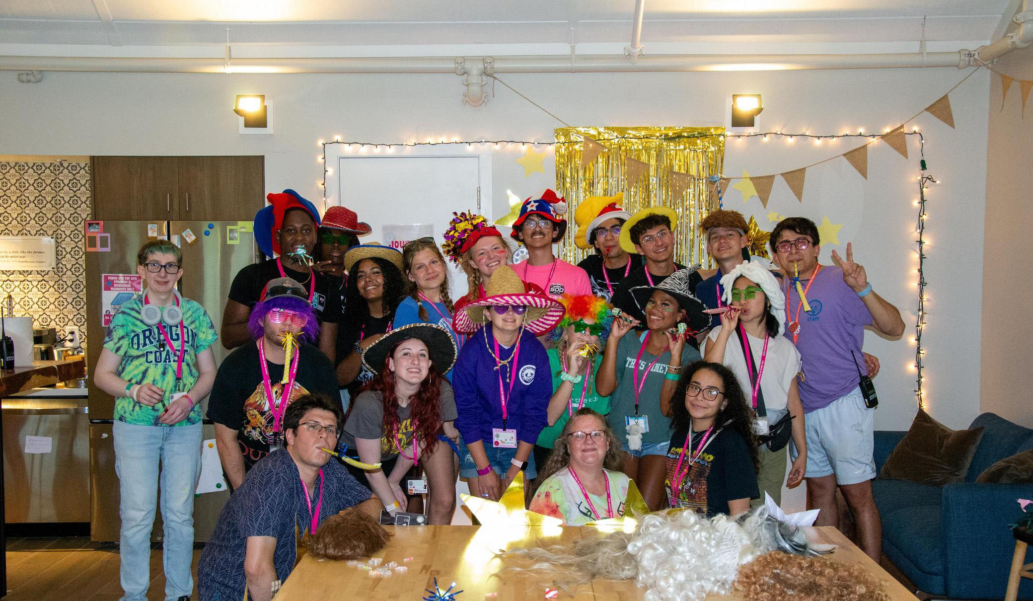 A group of people in colorful hats and costumes pose together in a decorated room with string lights and gold streamers, smiling and making peace signs.