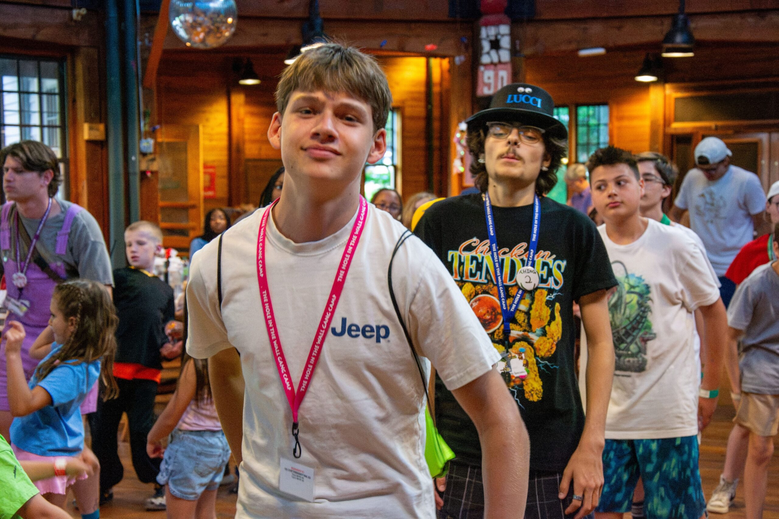 A group of children and teens dance indoors at a summer camp. The boy in front wears a white Jeep T-shirt and pink lanyard; others behind him wear colorful shirts and hats. The atmosphere is lively and cheerful.
