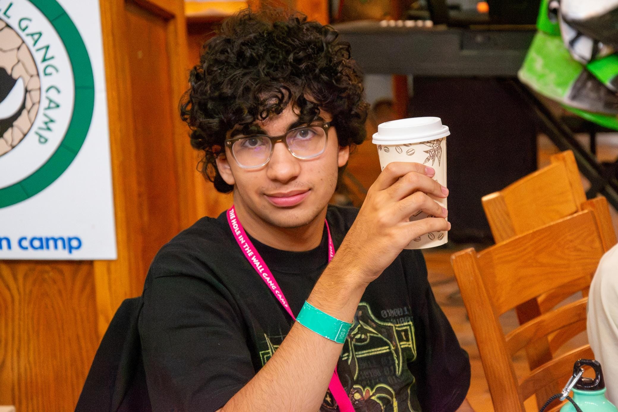 A person with curly hair and glasses holds a paper coffee cup, sitting at a table with wooden chairs. They wear a black shirt, pink lanyard, and teal wristband. A camp sign is partially visible in the background.
