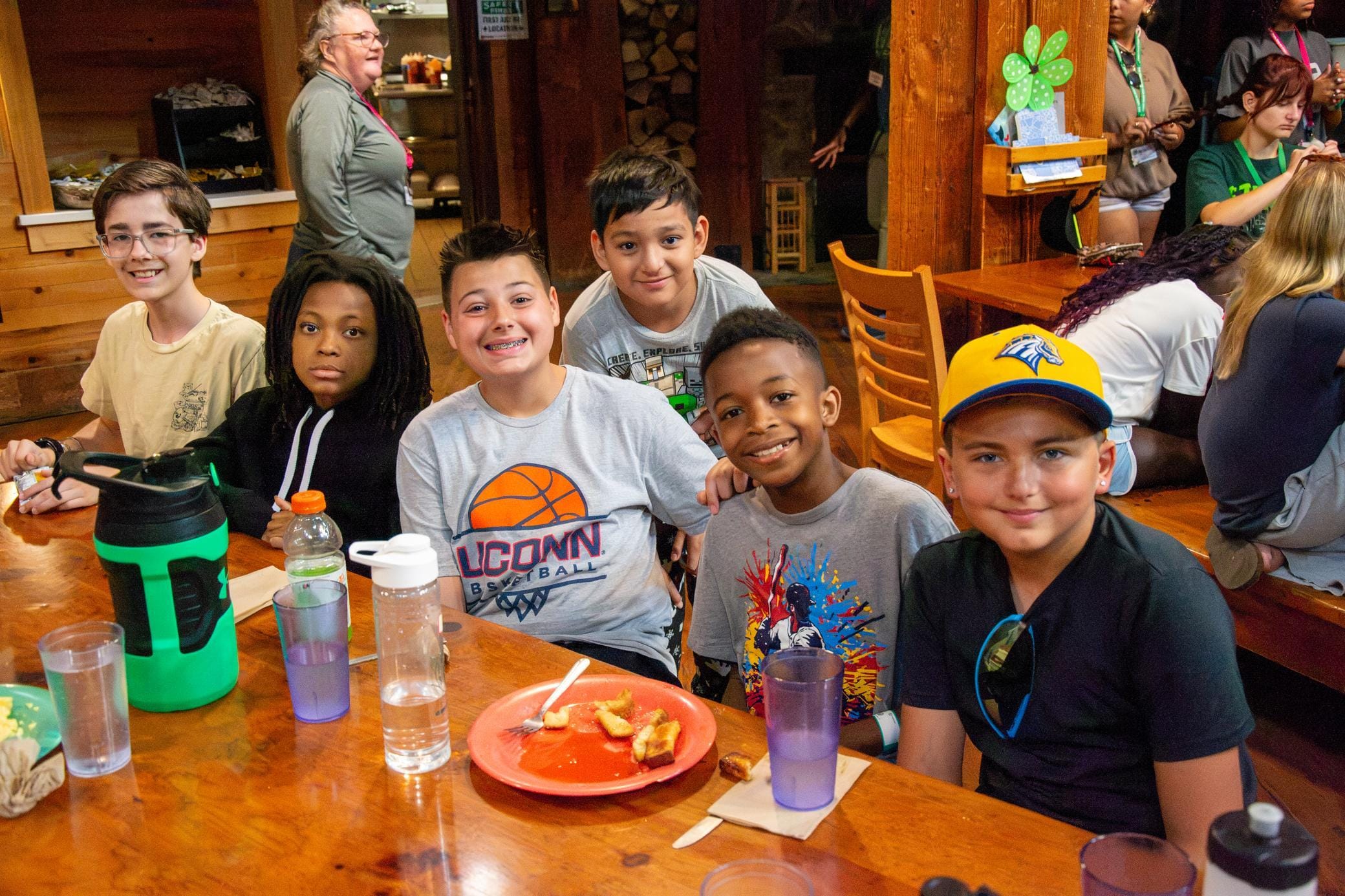 Six boys sit smiling at a wooden table in a cozy dining hall, with plates of food, water bottles, and cups around them. Other people are in the background enjoying the meal.