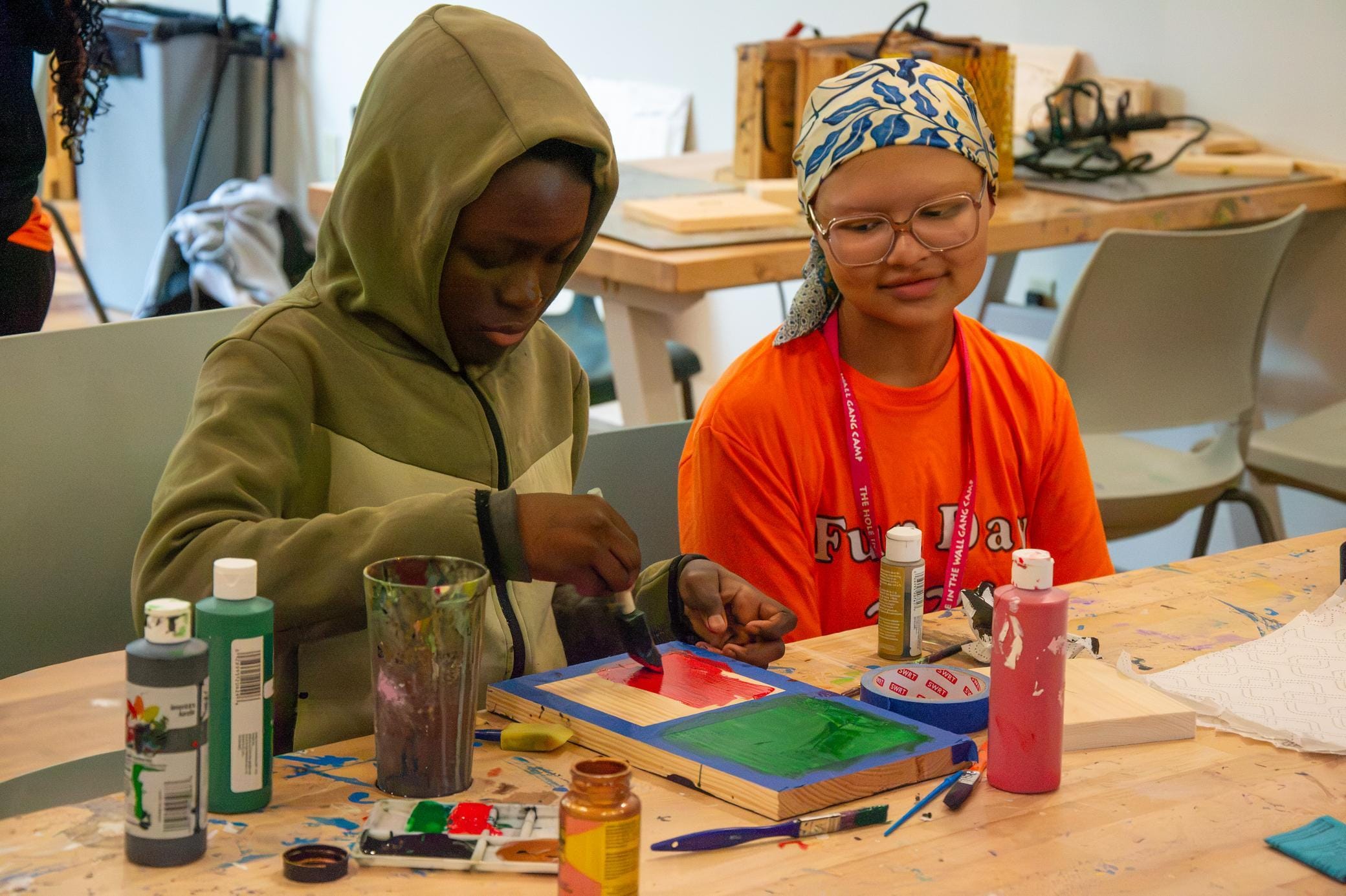 Two kids sit at a table painting wooden boards with bright colors. One wears a green hoodie and paints, while the other in an orange shirt and headscarf watches and smiles. Art supplies are scattered on the table.