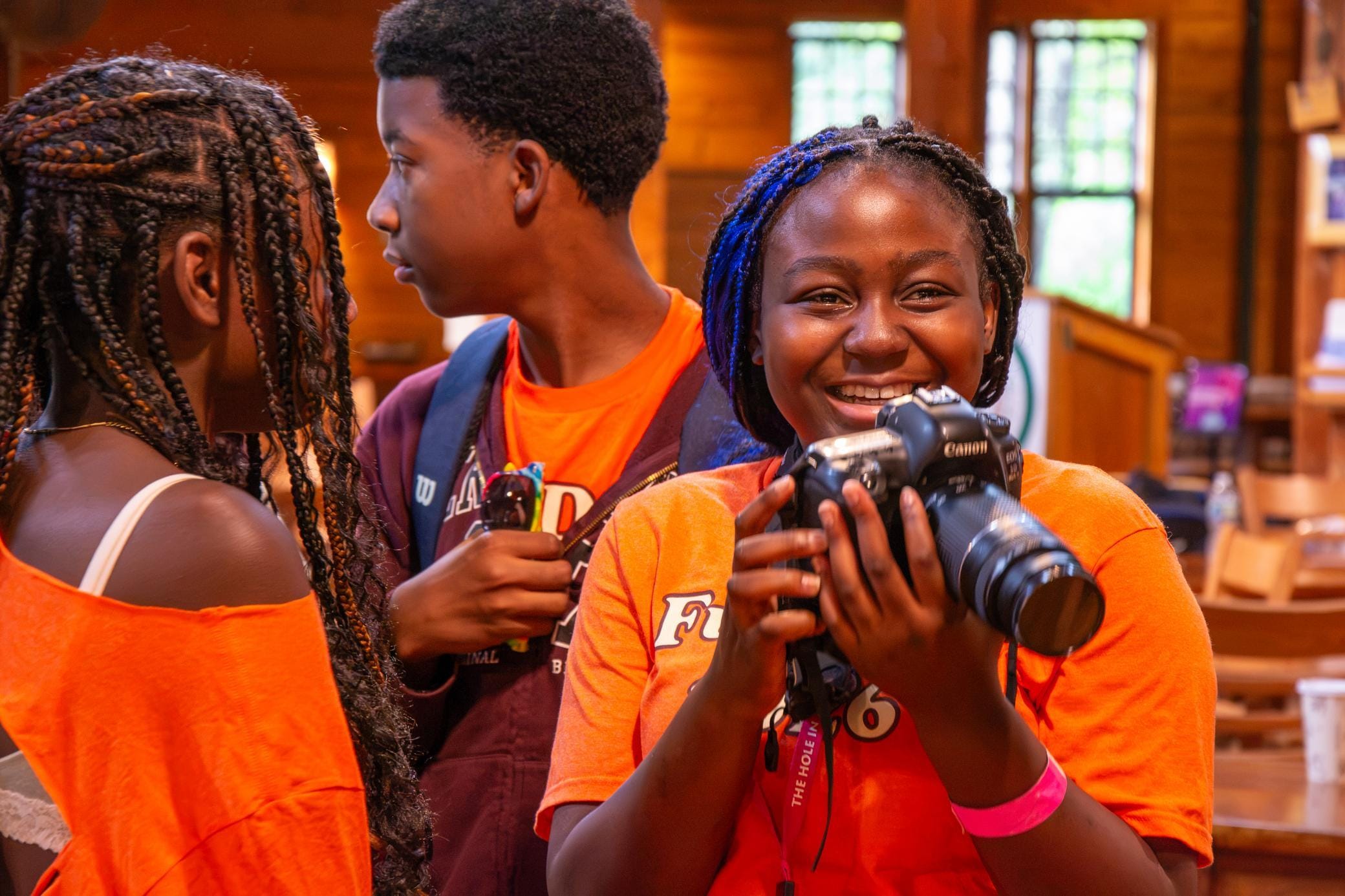 A smiling girl holds a camera, standing with two other kids indoors. All three wear orange shirts, and the background shows a wooden room with shelves and windows.
