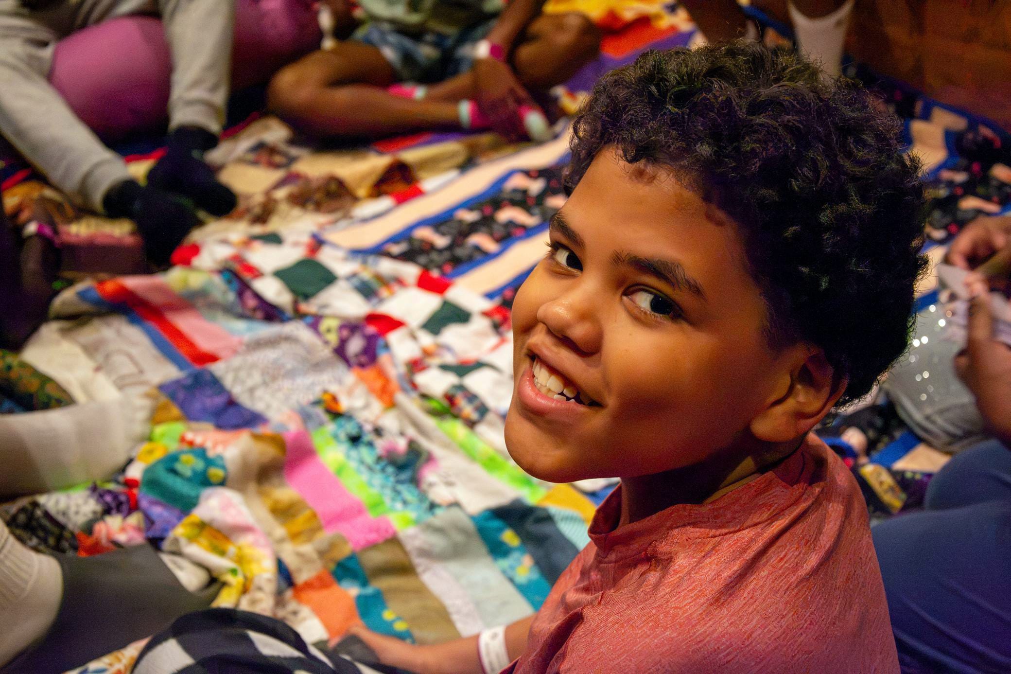A smiling child with curly hair looks over their shoulder while sitting on a colorful patchwork quilt, surrounded by others whose faces are out of frame.