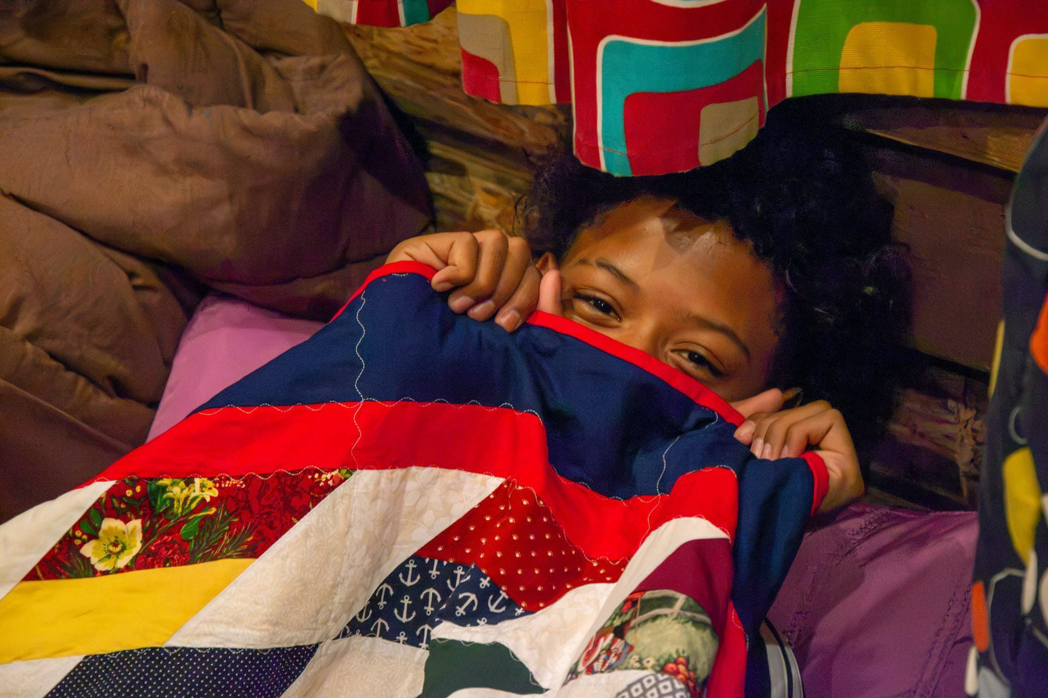 A child lies in bed, partially hidden under a colorful patchwork quilt, peeking out with their eyes visible and holding the quilt up near their face.