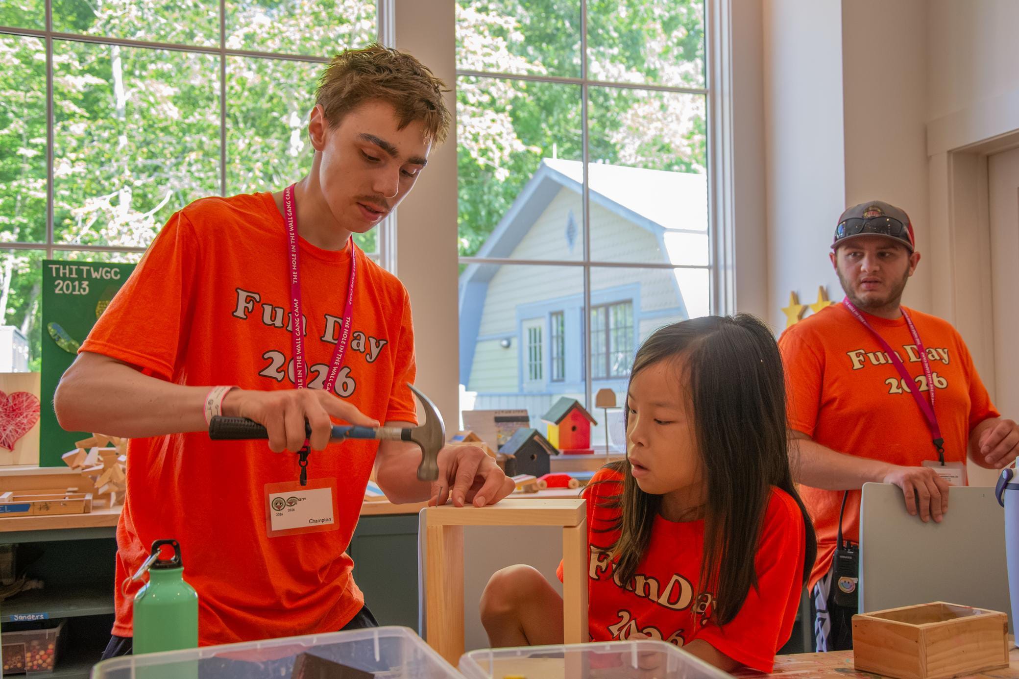 Three people in matching orange Fun Day 2026 shirts work on a wood project indoors; a young man hammers while a girl watches closely and another man looks on in the background.
