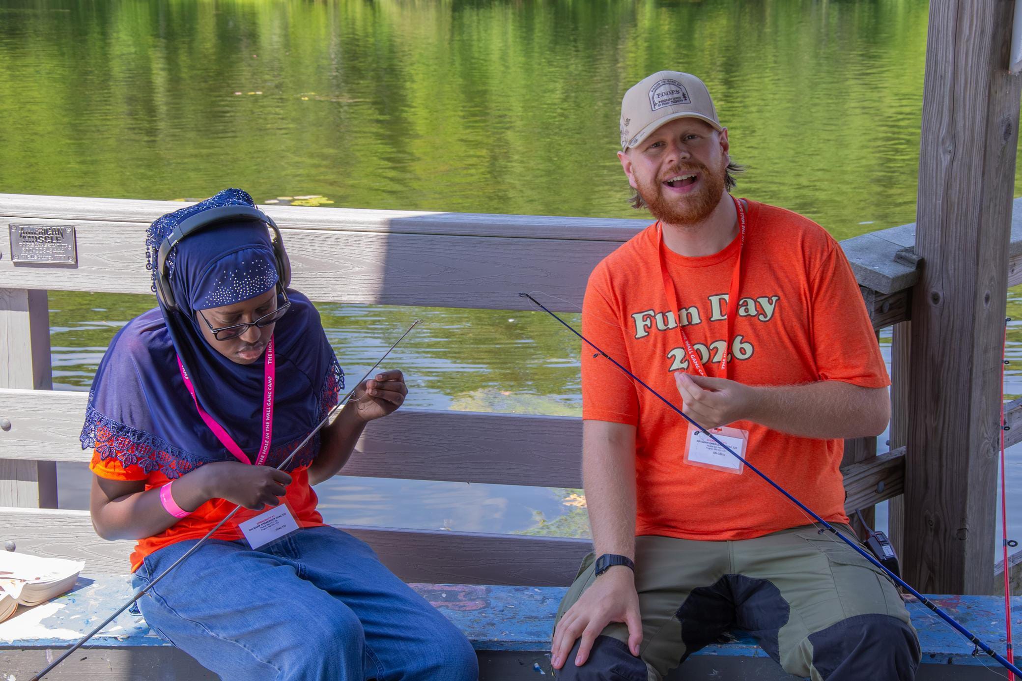 Two people sit on a dock by the water, fishing. One adjusts a fishing line, while the other, wearing a Fun Day 2026 shirt, smiles at the camera. Trees and water are visible in the background.