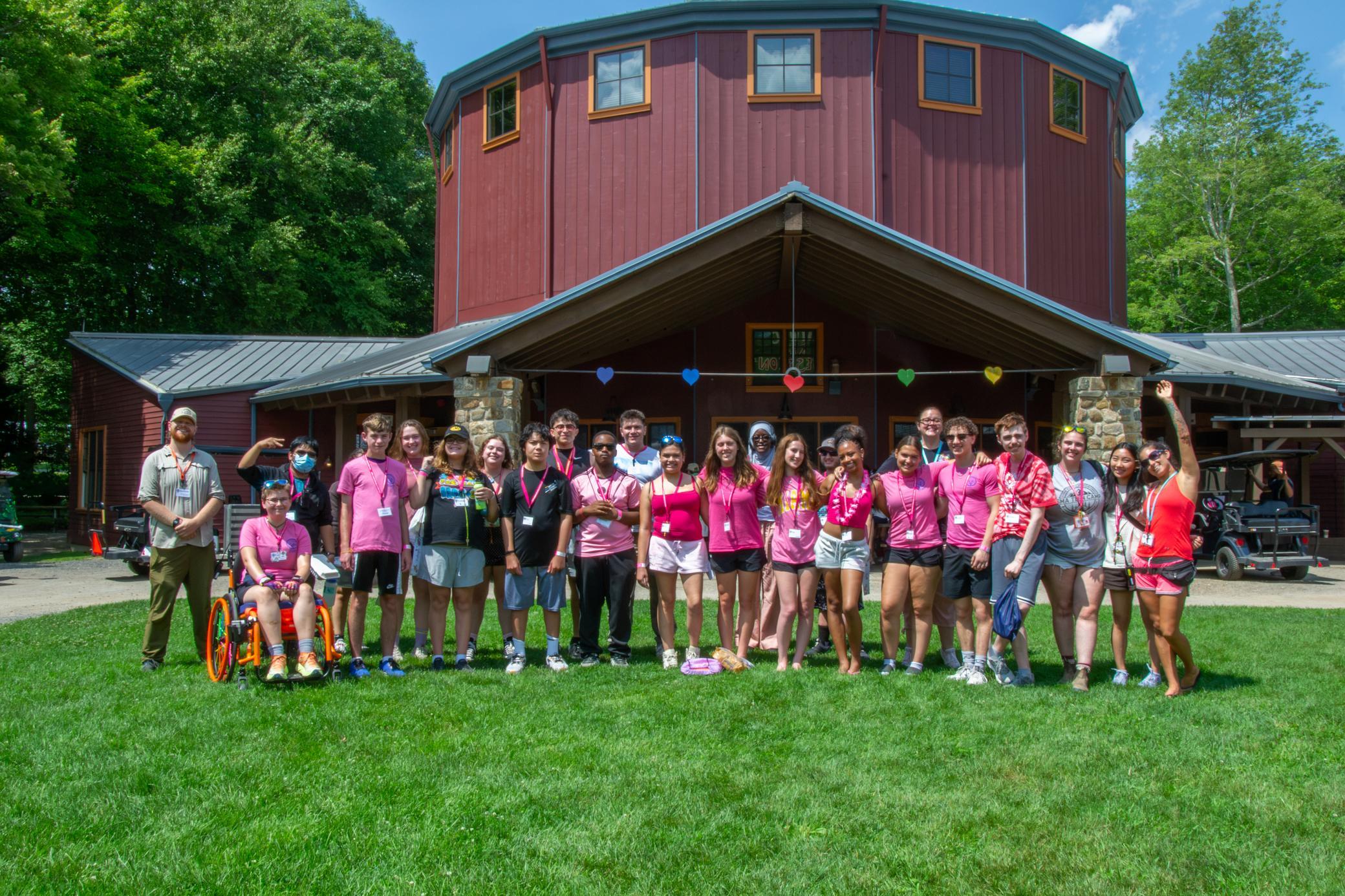 A diverse group of people, some in pink shirts, stand and pose for a photo on a lawn in front of a red building with heart decorations and trees in the background.
