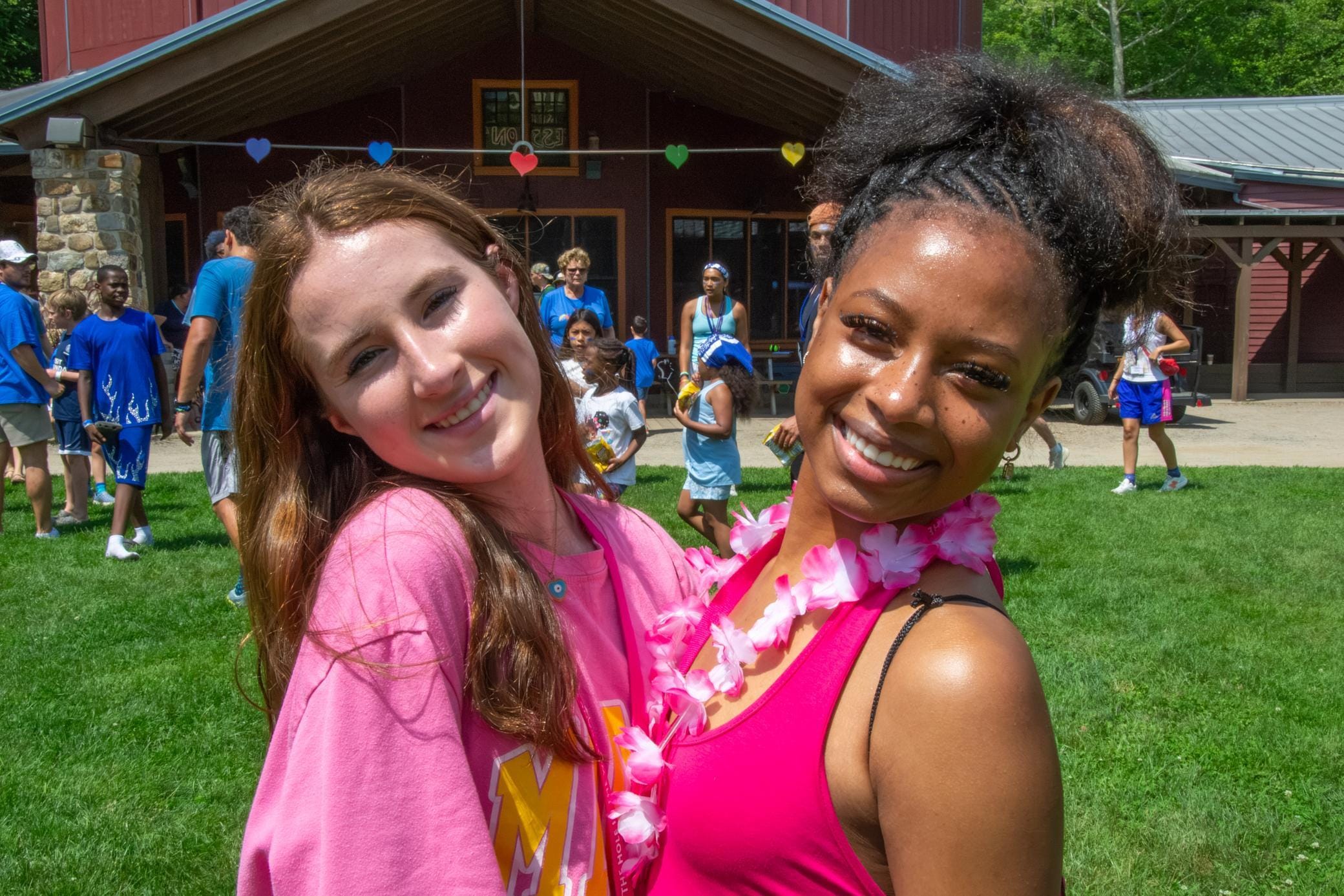 Two girls smiling and posing together outside at a summer camp, with other campers and counselors in the background. One girl wears a pink shirt, the other a pink tank top and a flower lei. Bright, sunny day.