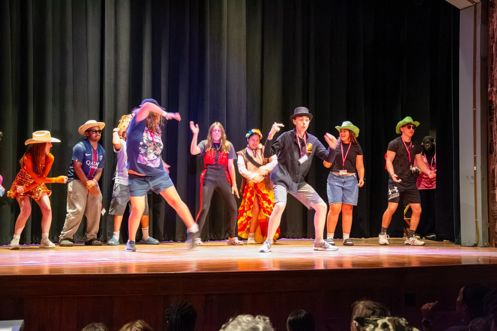 A group of people in colorful hats and costumes dance energetically on a brightly lit stage with a dark curtain backdrop.