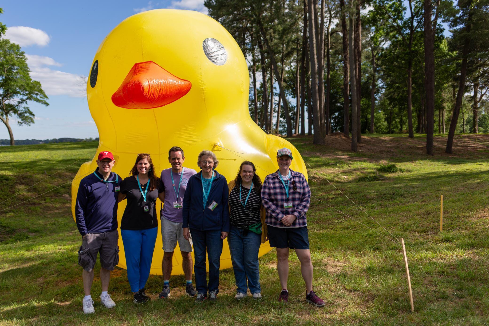 Seven people stand smiling in front of a large inflatable yellow duck outdoors on a sunny day with trees and grass in the background.