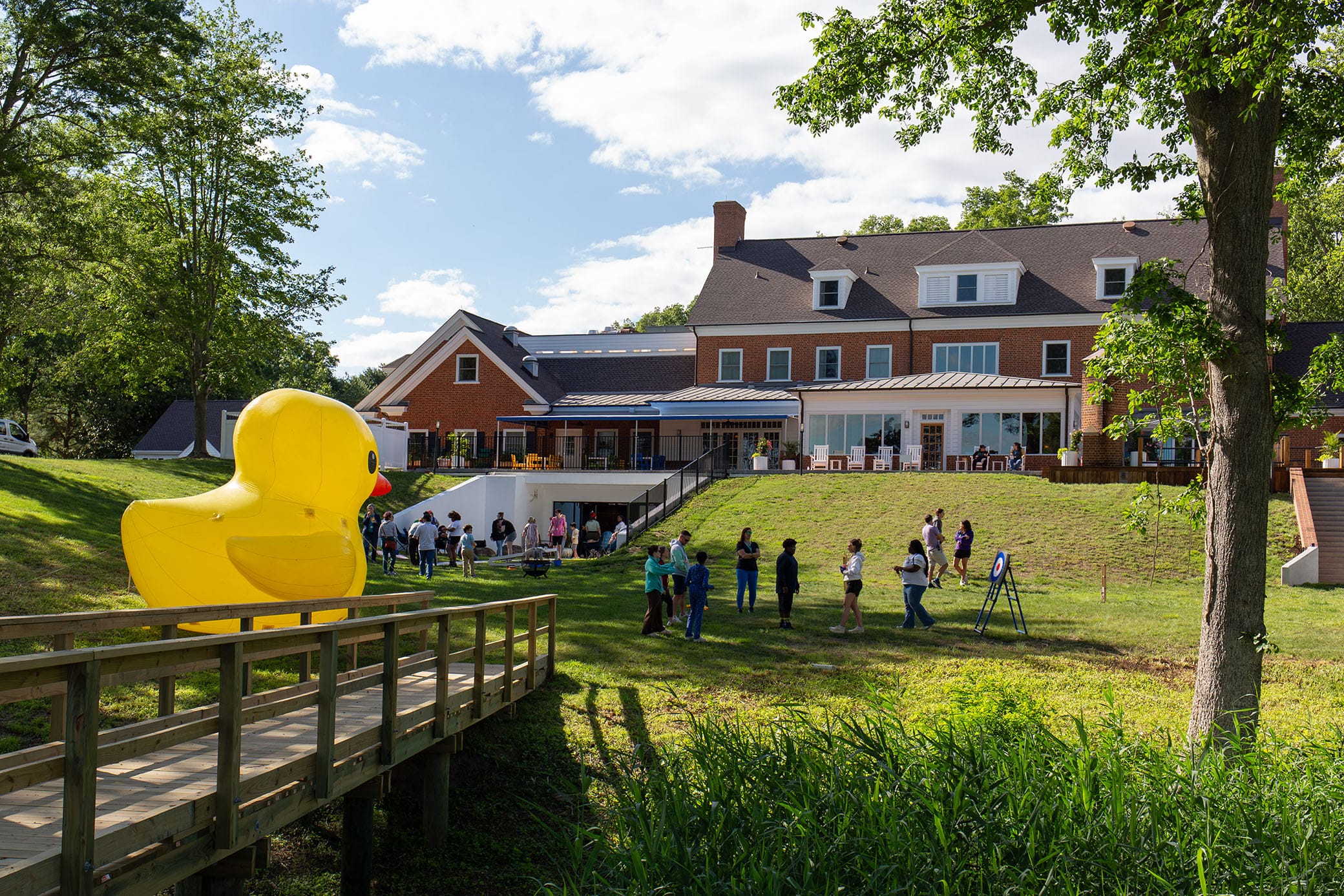 A group of people stands in line on a lawn near a large brick building; a giant yellow rubber duck sculpture is visible on a wooden walkway in the foreground. Trees and blue sky frame the scene.
