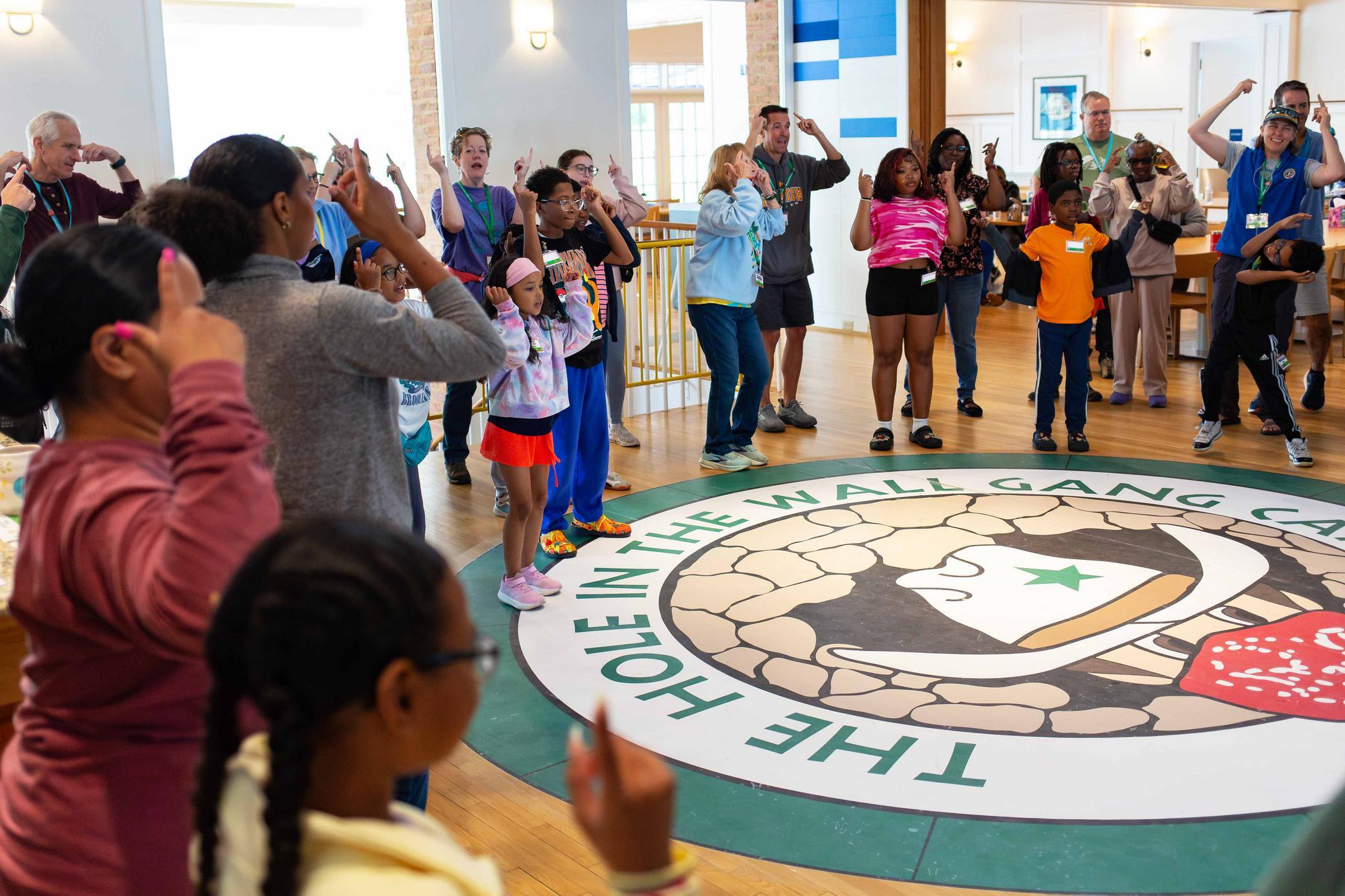 A diverse group of children and adults stand in a circle, following instructions and making hand gestures on a wooden floor with a large camp logo that says The Hole in the Wall Gang Camp..
