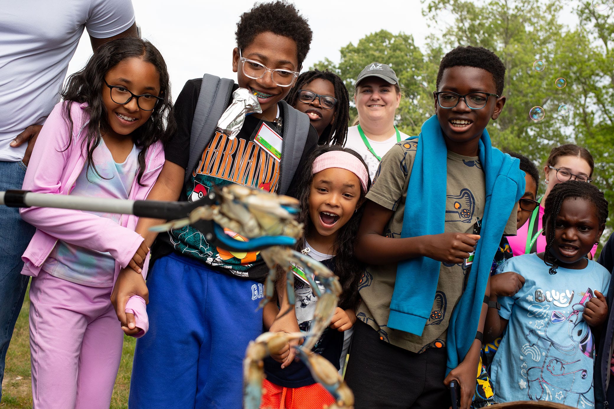 A group of children and adults smile and laugh outdoors, watching bubbles being made. Some kids wear glasses and backpacks, and the scene is lively with trees in the background.