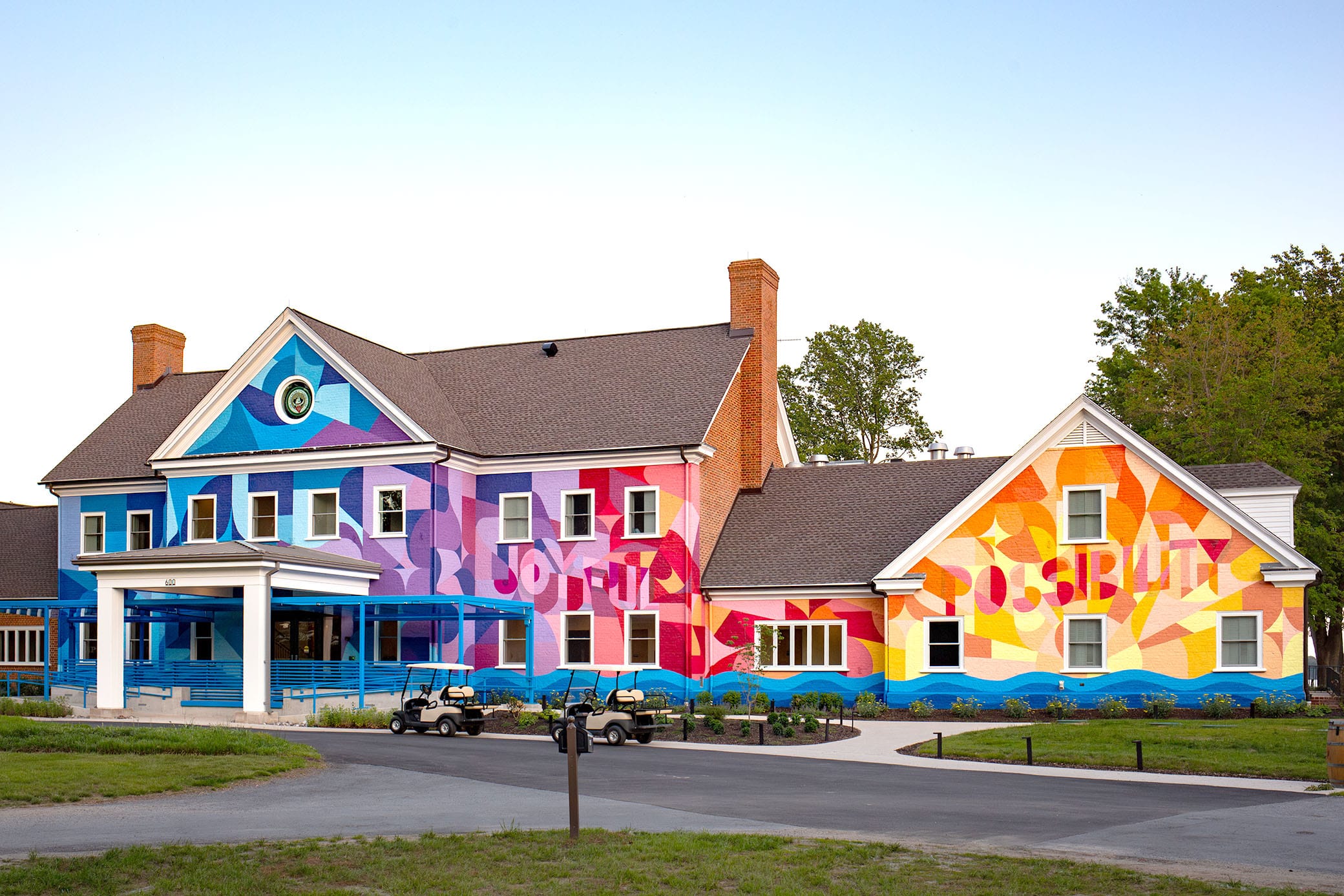 A large building with a vibrant, colorful mural featuring abstract shapes and the word possibility painted across its facade, surrounded by grass and trees, with two golf carts parked out front.
