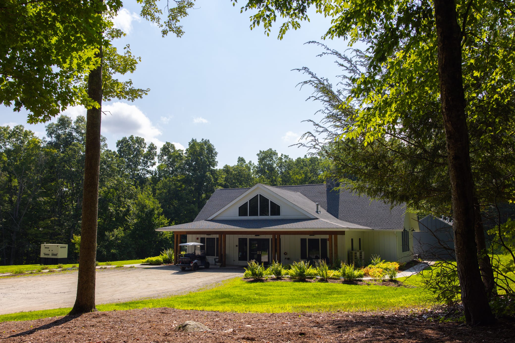 A house with a large porch sits among green trees on a sunny day. A golf cart is parked outside, and a well-kept lawn surrounds the building. The sky is clear with a few clouds visible.