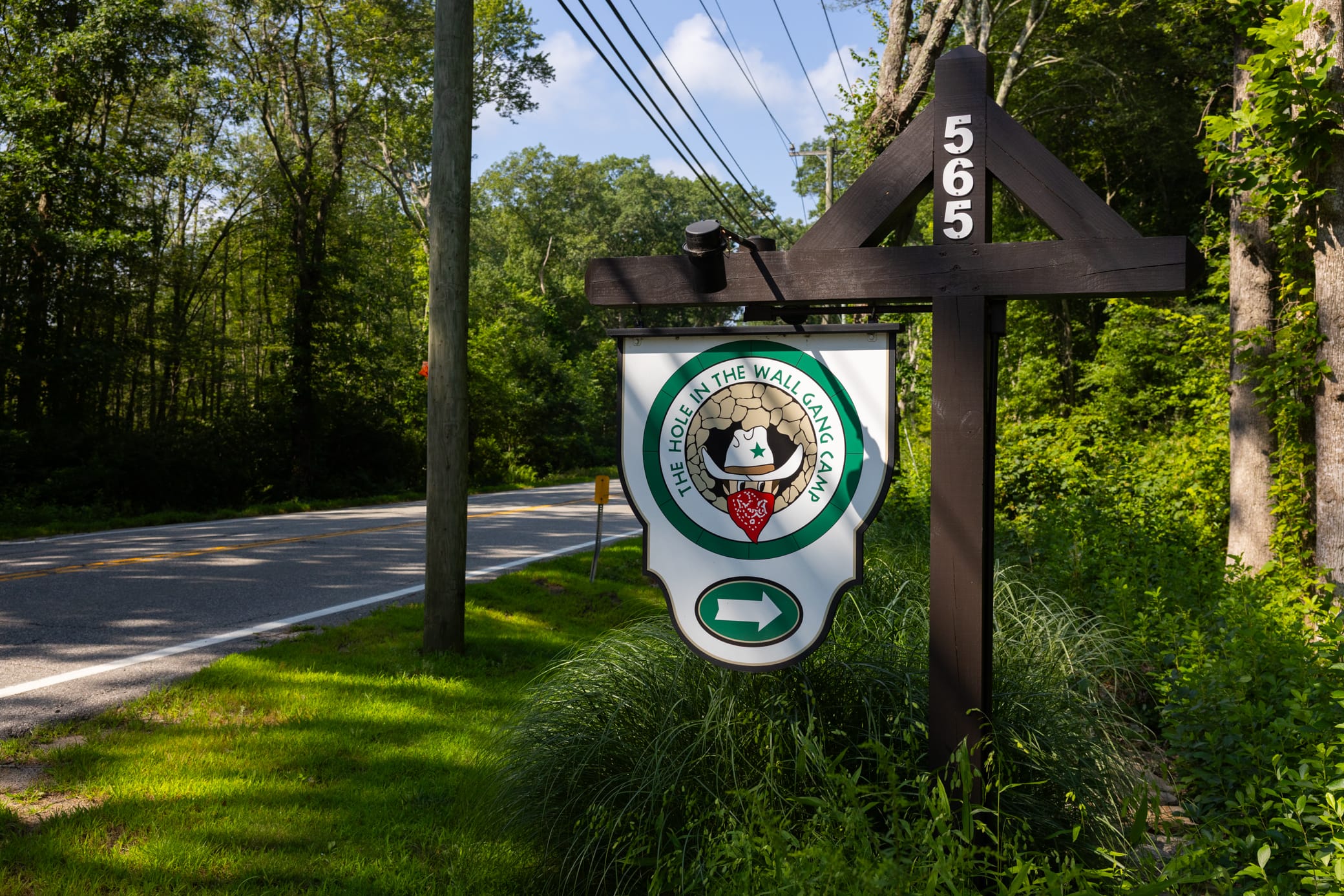 A roadside sign at 565 marks the entrance to The Hole in the Wall Gang Camp, featuring the camps logo and a right arrow. Trees and a telephone pole line the road in the background.