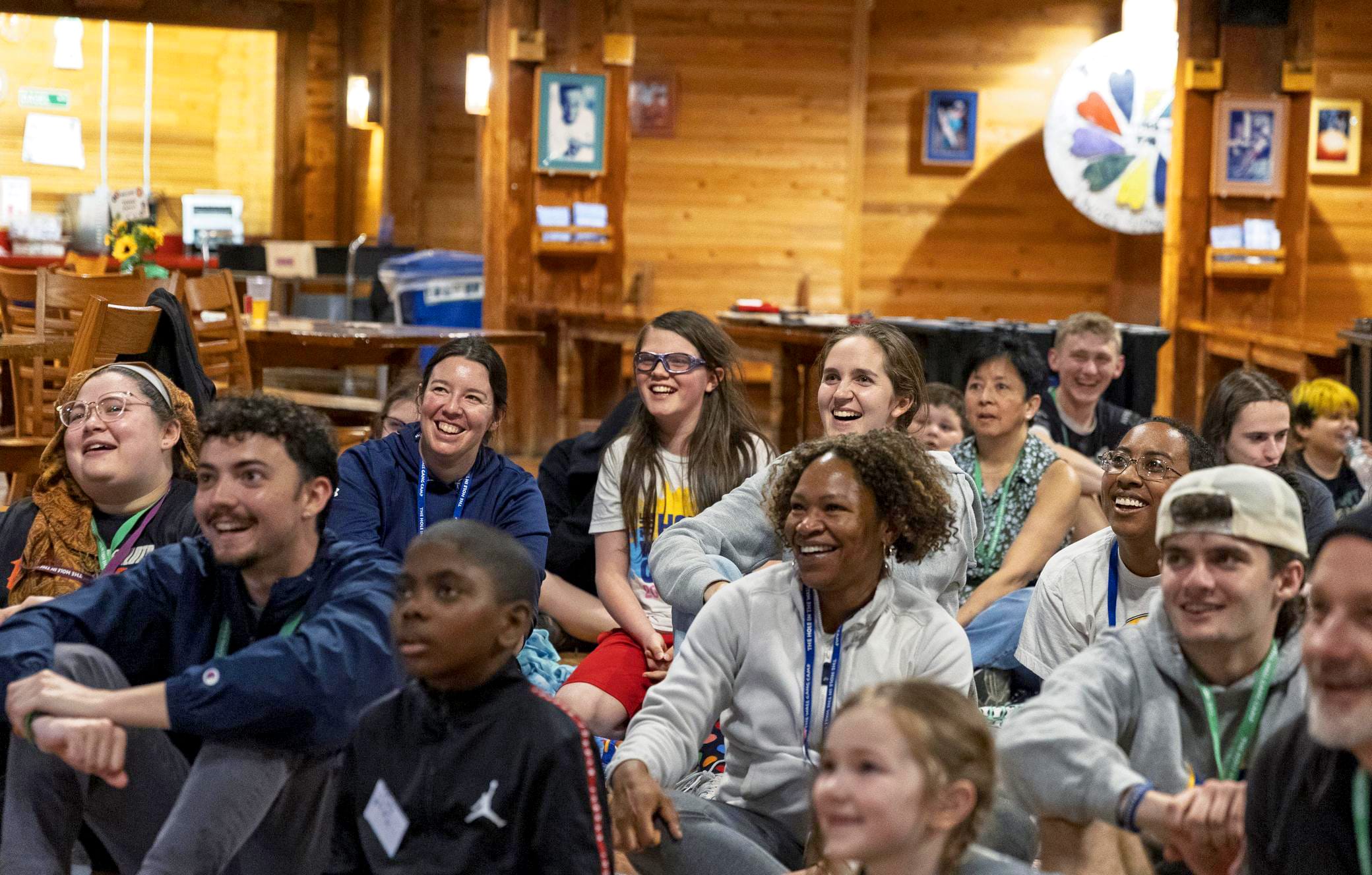 A diverse group of people, both adults and children, sit closely together indoors, smiling and laughing as they watch something off-camera in a cozy, wooden room.
