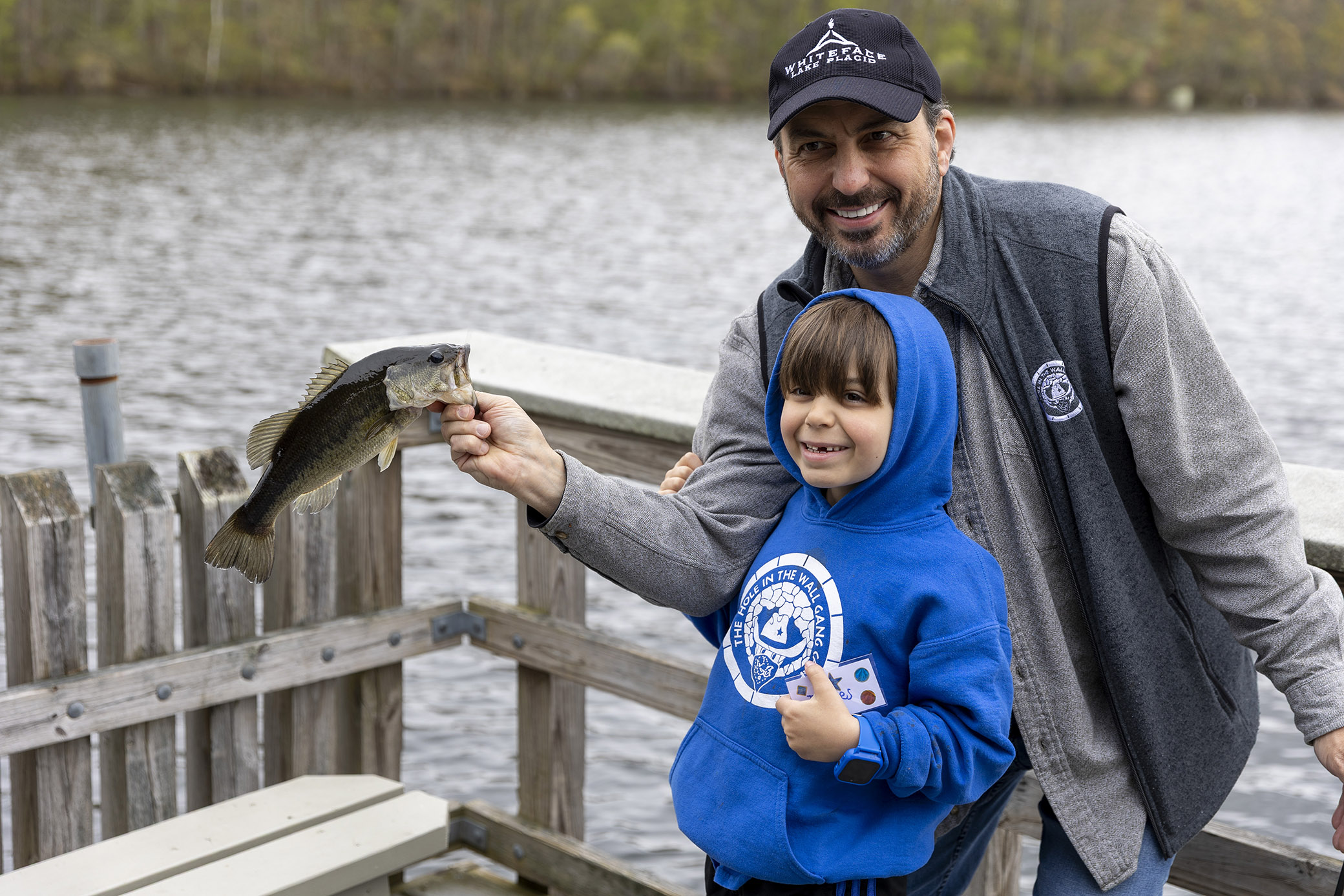 A man and a child smile by a lakeside dock, proudly holding up a fish they caught together. The child wears a blue hoodie, and the man wears a cap and gray jacket. Trees and water are visible in the background.