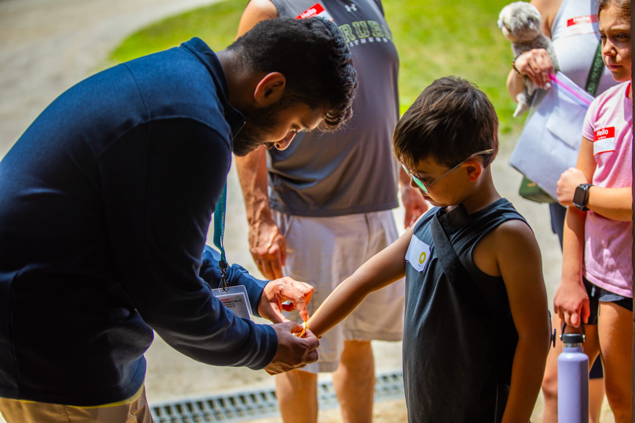 A man ties a wristband on a young boy outdoors, while other people, including a person holding a dog, watch nearby.