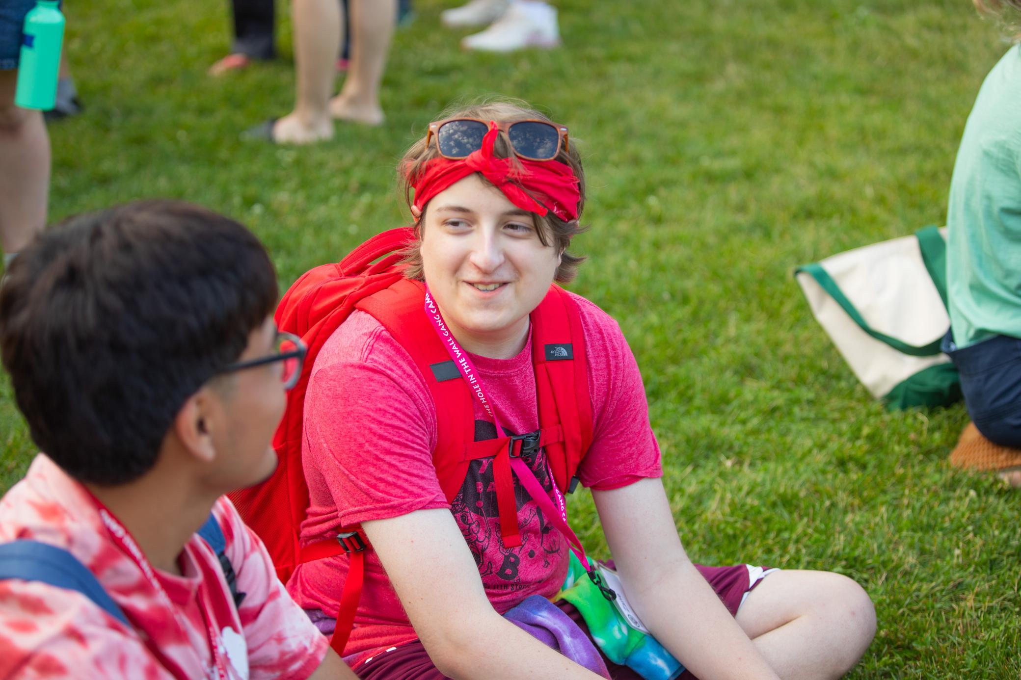 Two people sit on green grass. The person in front wears a red headband, sunglasses, a red backpack, and a pink shirt. They are smiling at the person beside them, who is mostly out of focus. Other people are seen in the background.