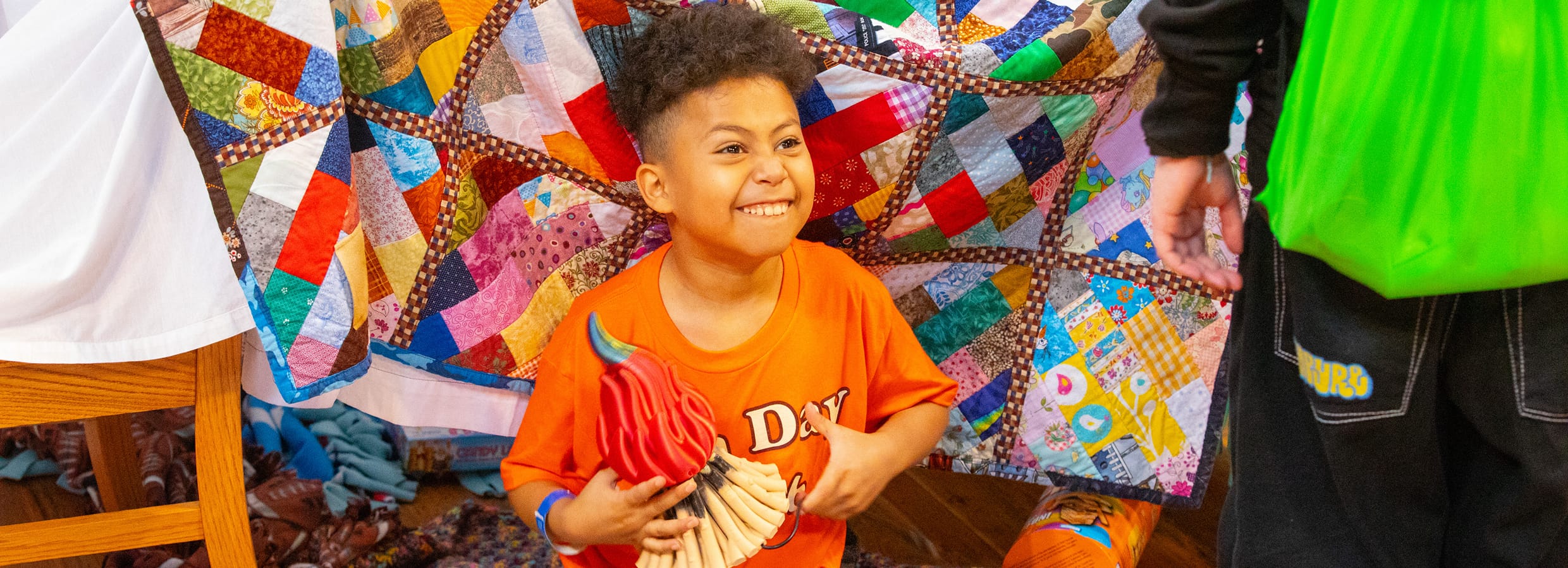 Smiling child holding a colorful wooden rooster sits in front of a patchwork quilt, wearing an orange shirt, next to an adult carrying a bright green bag.