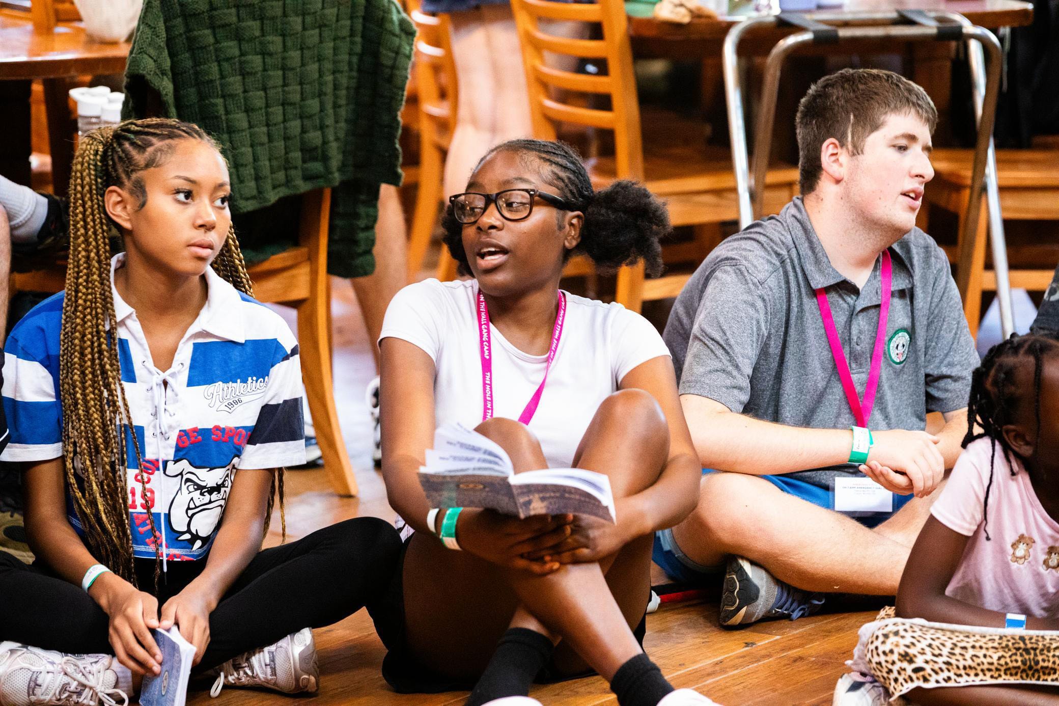 Three teens sit on a wooden floor in a lively indoor setting, holding books and wearing lanyards, while talking and listening. Wooden chairs and tables are visible in the background.
