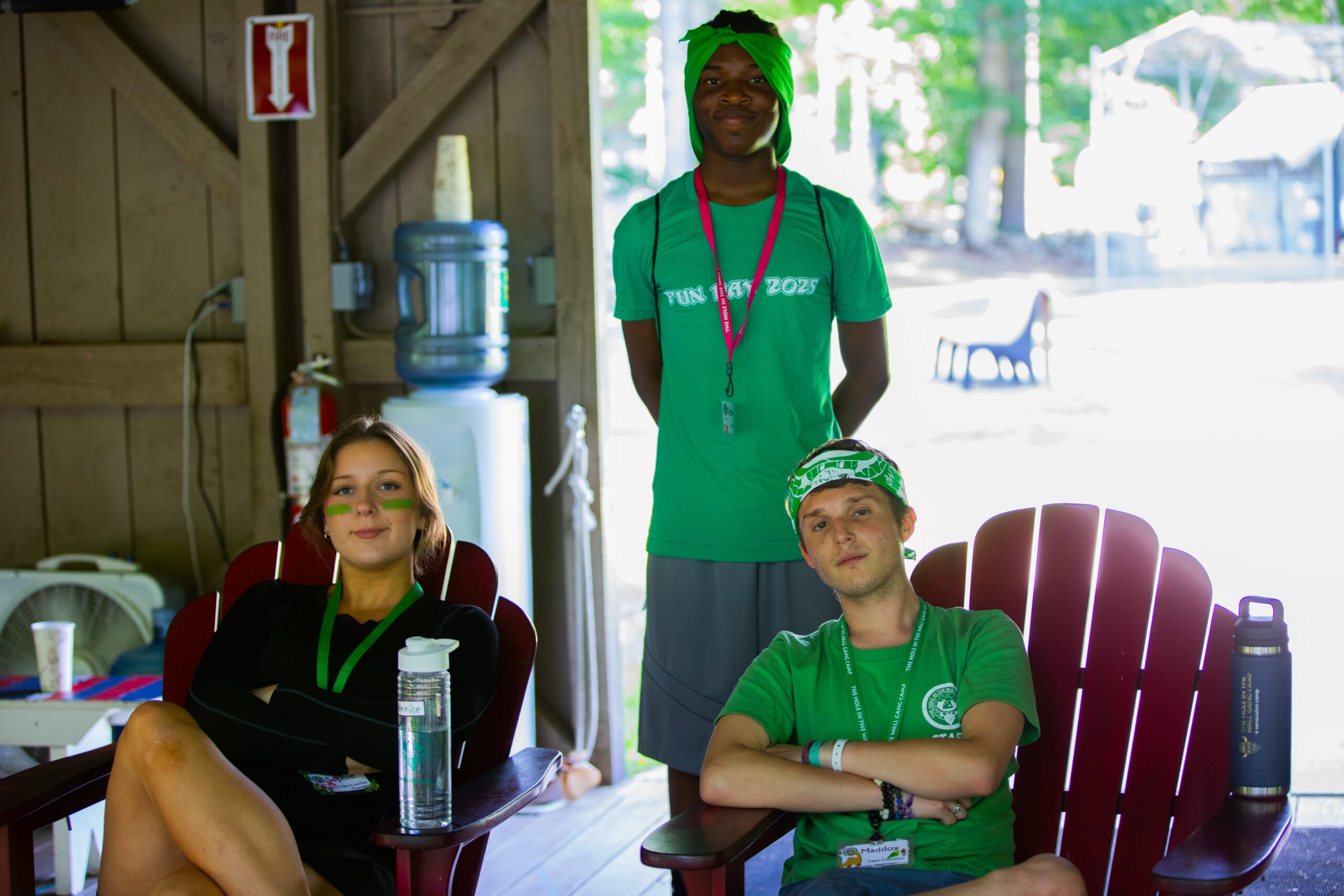 Three people pose indoors at camp; two sit on red chairs, one stands behind. All wear green lanyards. The standing person and one seated wear green shirts. A water cooler, cup, and trees outside are visible in the background.