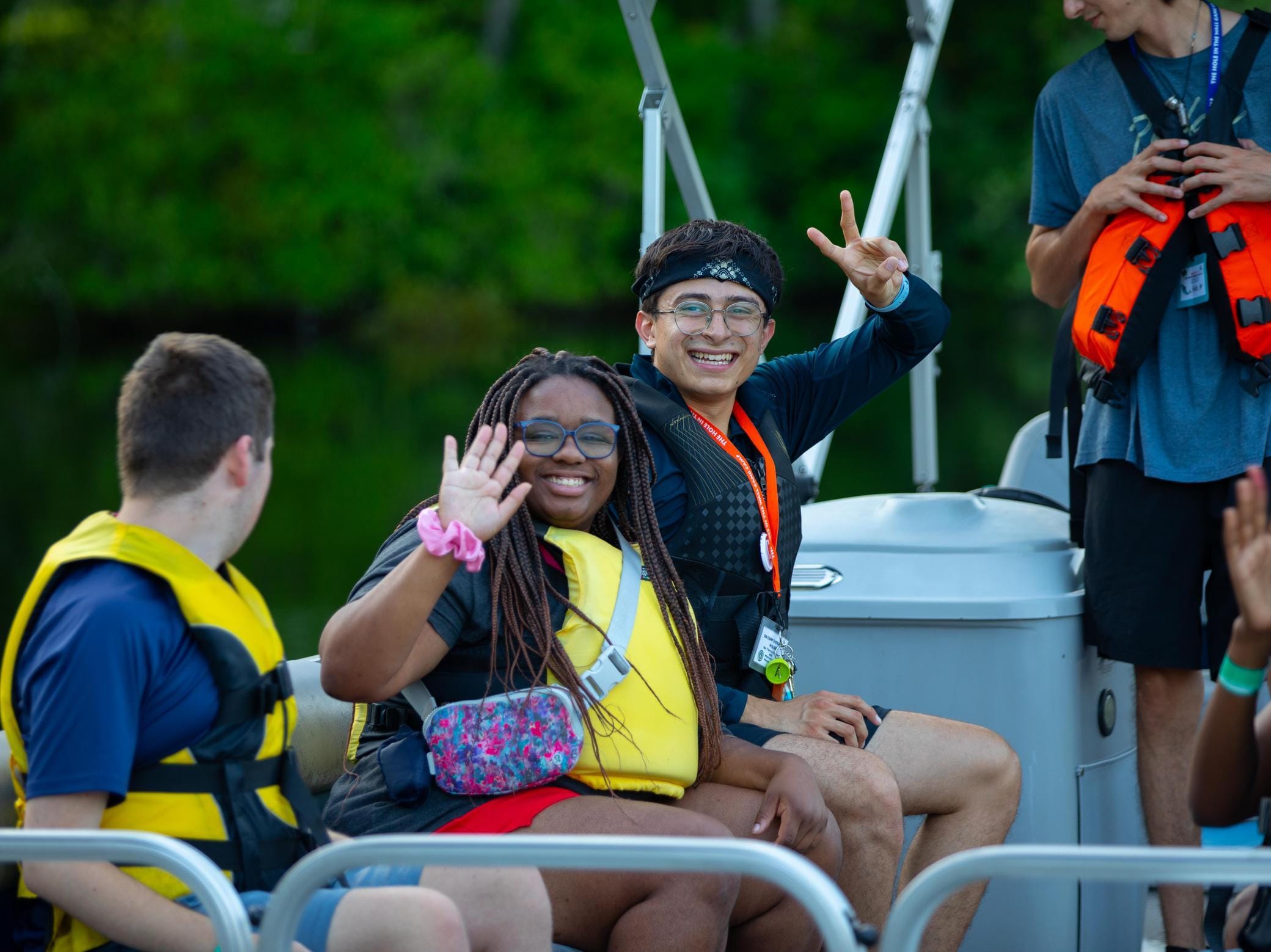 Four young people wearing life jackets sit on a boat. Two are smiling at the camera, one waving and the other making a peace sign. Trees and water are visible in the background.