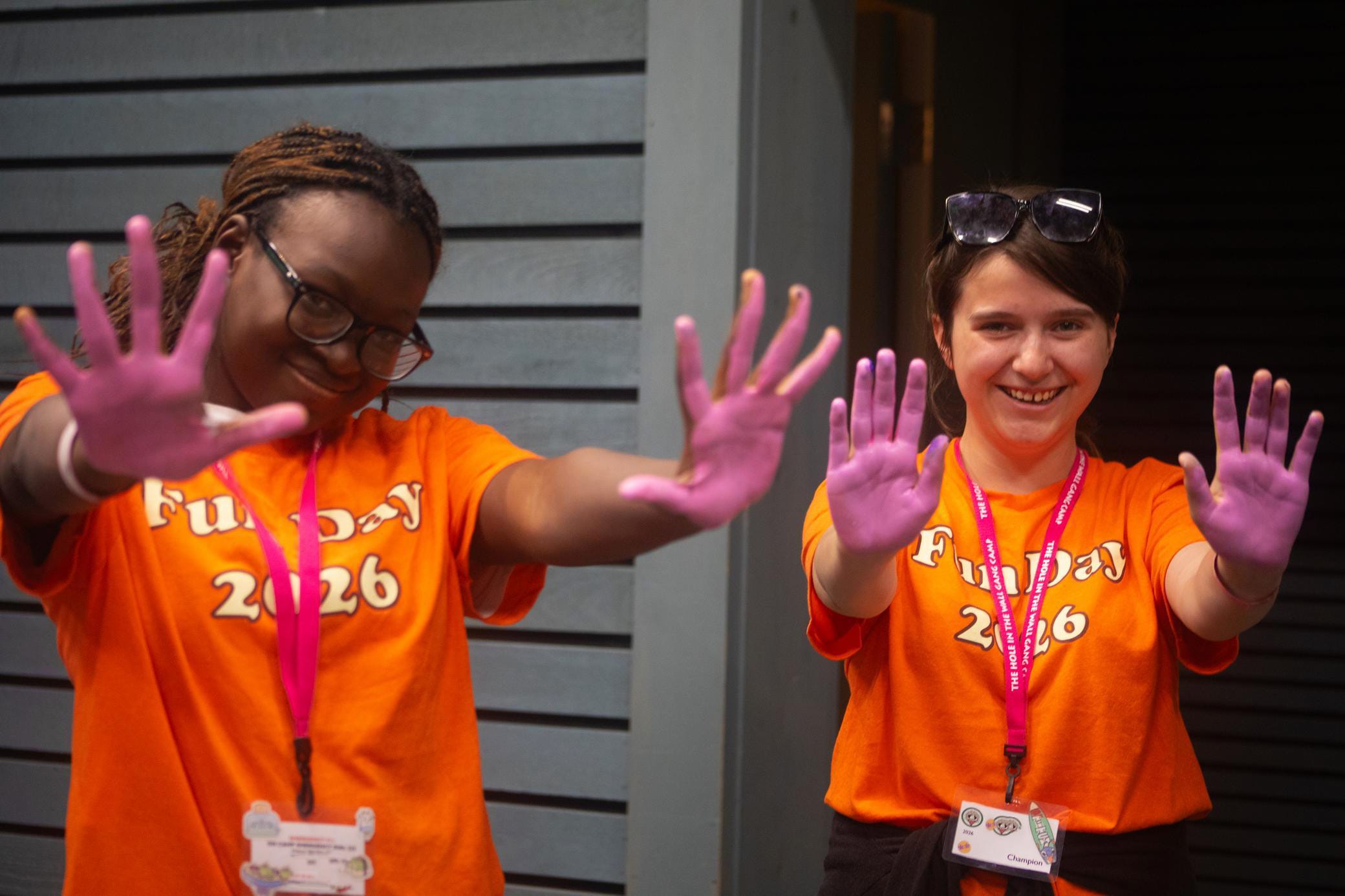 Two people in orange Funday 2026 shirts smile and show their hands, covered in purple paint, toward the camera. Both wear name tags and pink lanyards. They stand in front of a wooden wall.