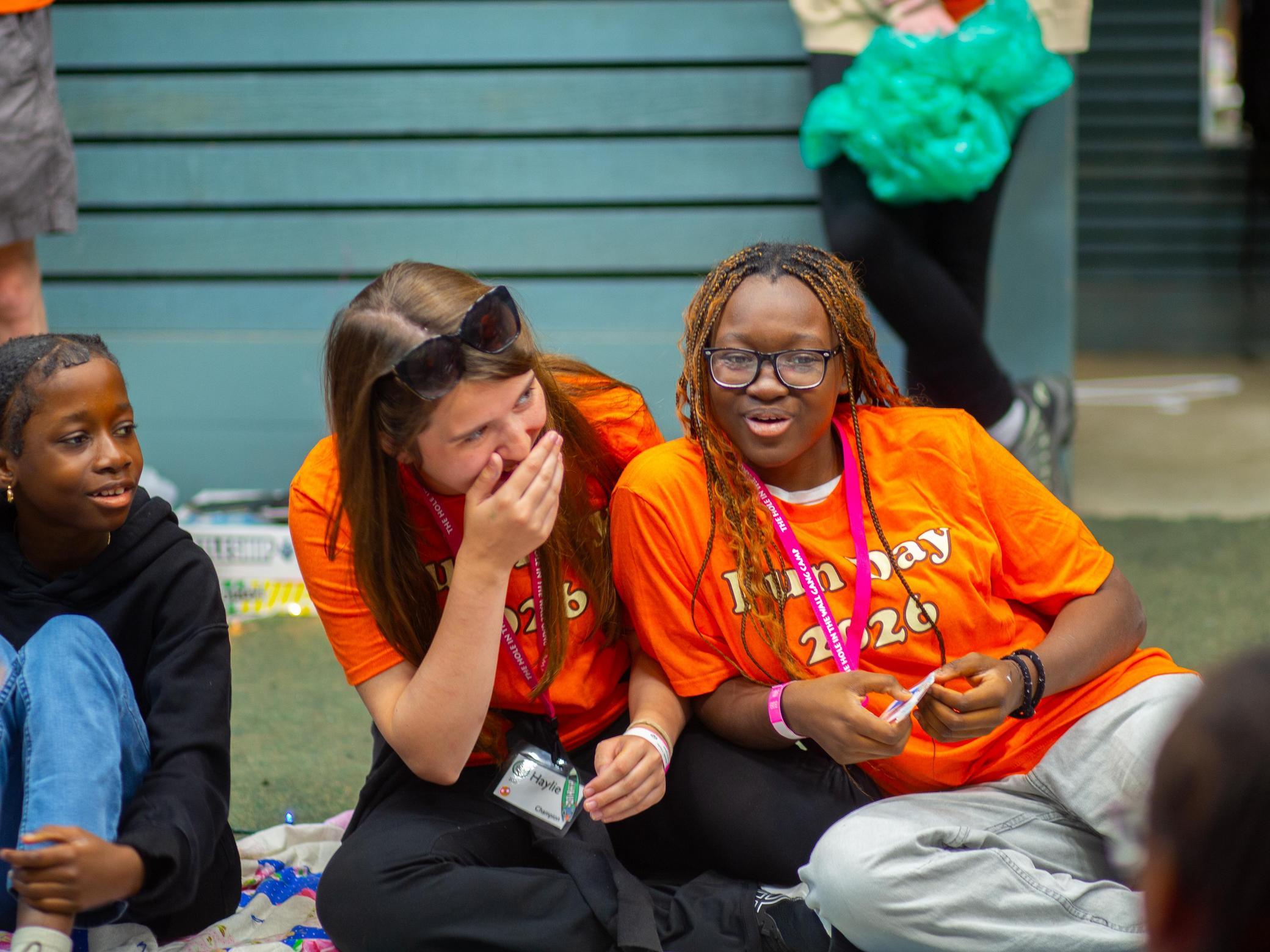 Three young people in bright orange Fun Day 2026 shirts sit on the ground, smiling and laughing together. Two of them are close, one covering her mouth while laughing. The background shows blurred people and a building wall.