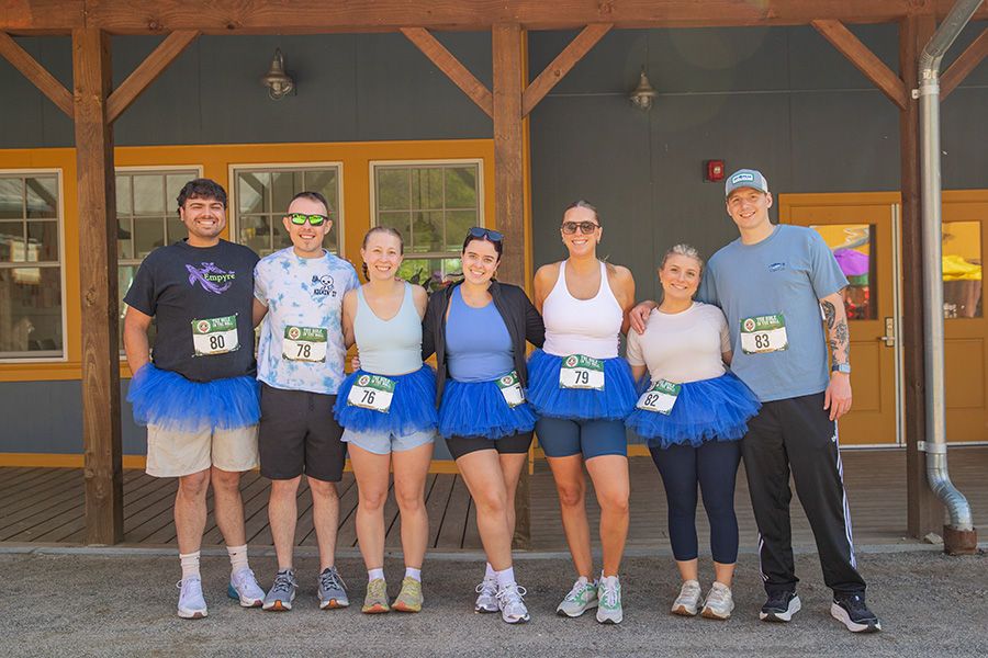 Seven people pose outside a building, six wearing blue tutus and athletic outfits with race bibs, smiling at the camera before a running event.