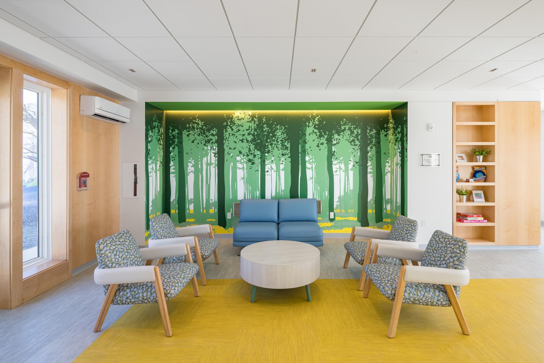 Modern waiting area with four patterned chairs, a blue loveseat, and a round table on a yellow rug. The back wall features a green forest mural, and wooden accents and natural light create a welcoming atmosphere.