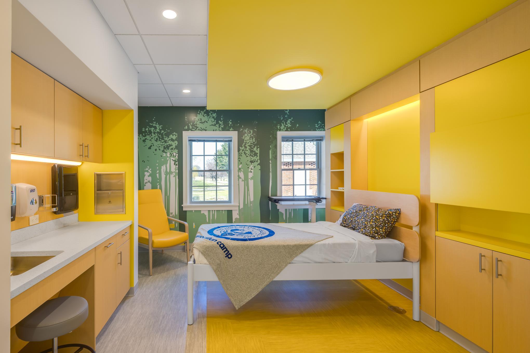 A modern hospital room with yellow accents, a single bed, a chair near two windows, built-in cabinets, a small sink area, and a blanket with a blue logo on the bed.