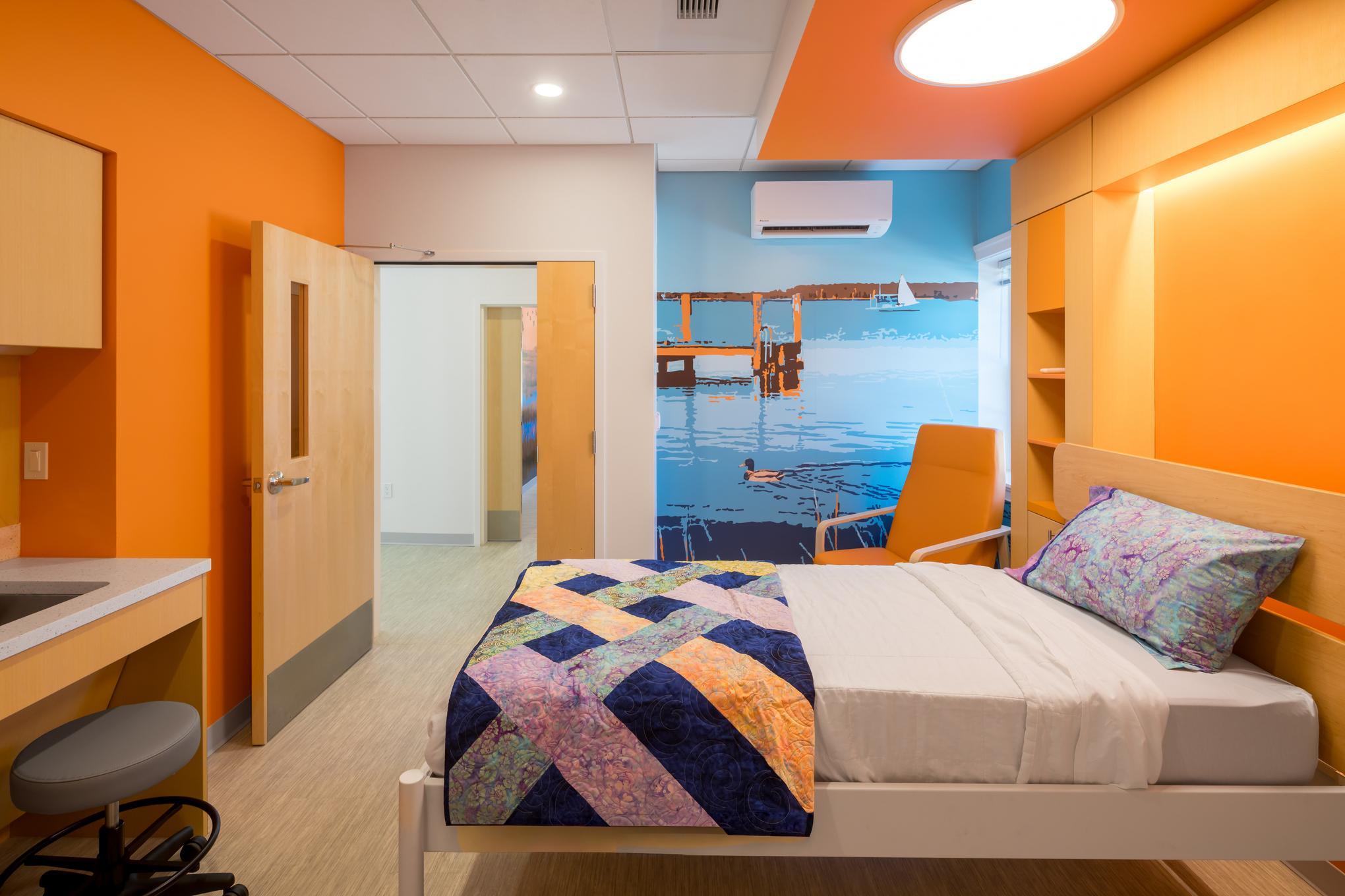 A brightly lit hospital room with orange walls, a single bed with a colorful quilt, a sink, chair, and a wall mural showing a dock and water scene.