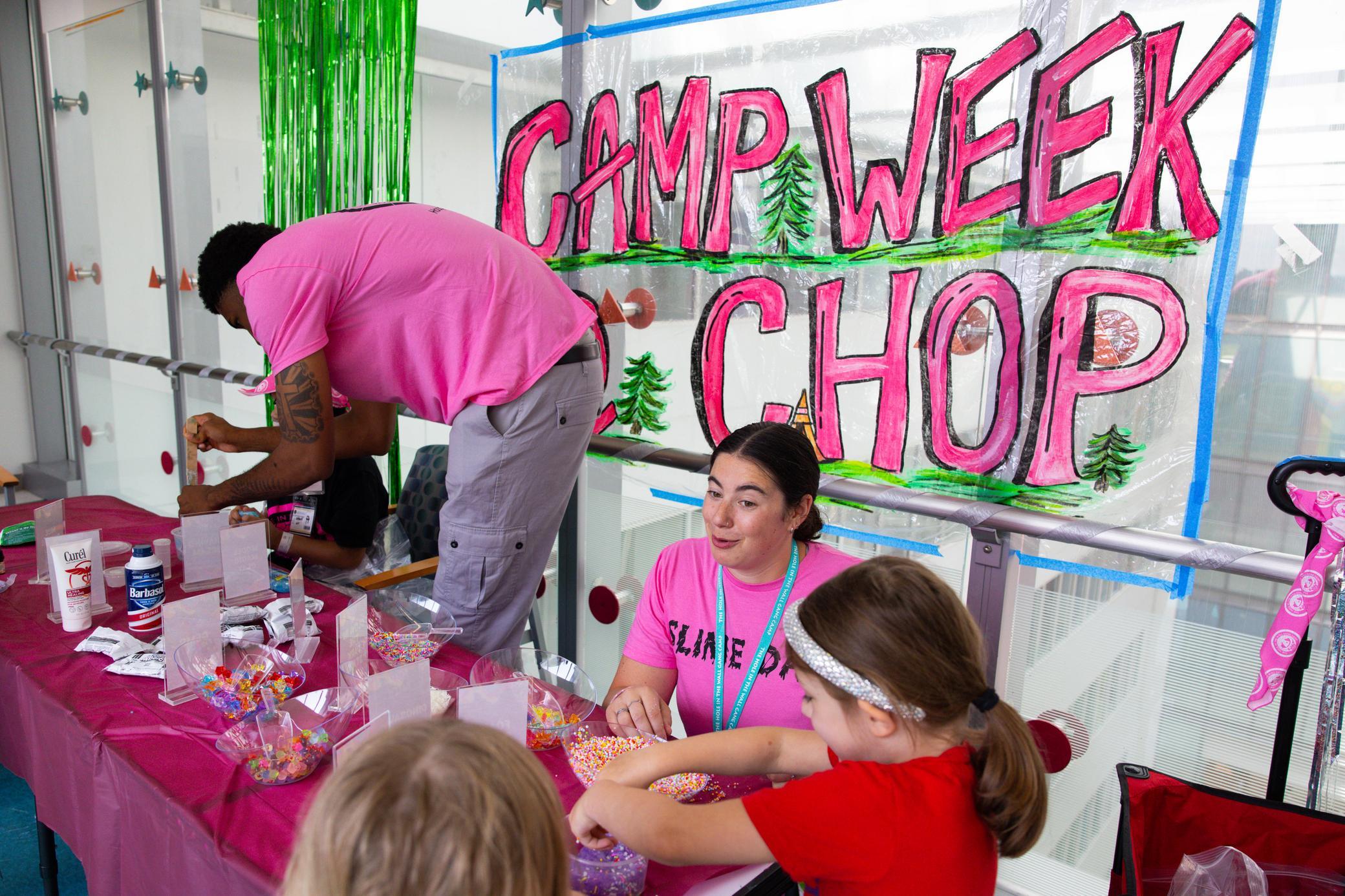 Children make crafts at a table with supplies, guided by two adults, under a colorful Camp Week CHOP sign decorated with trees. Everyone appears engaged and festive.