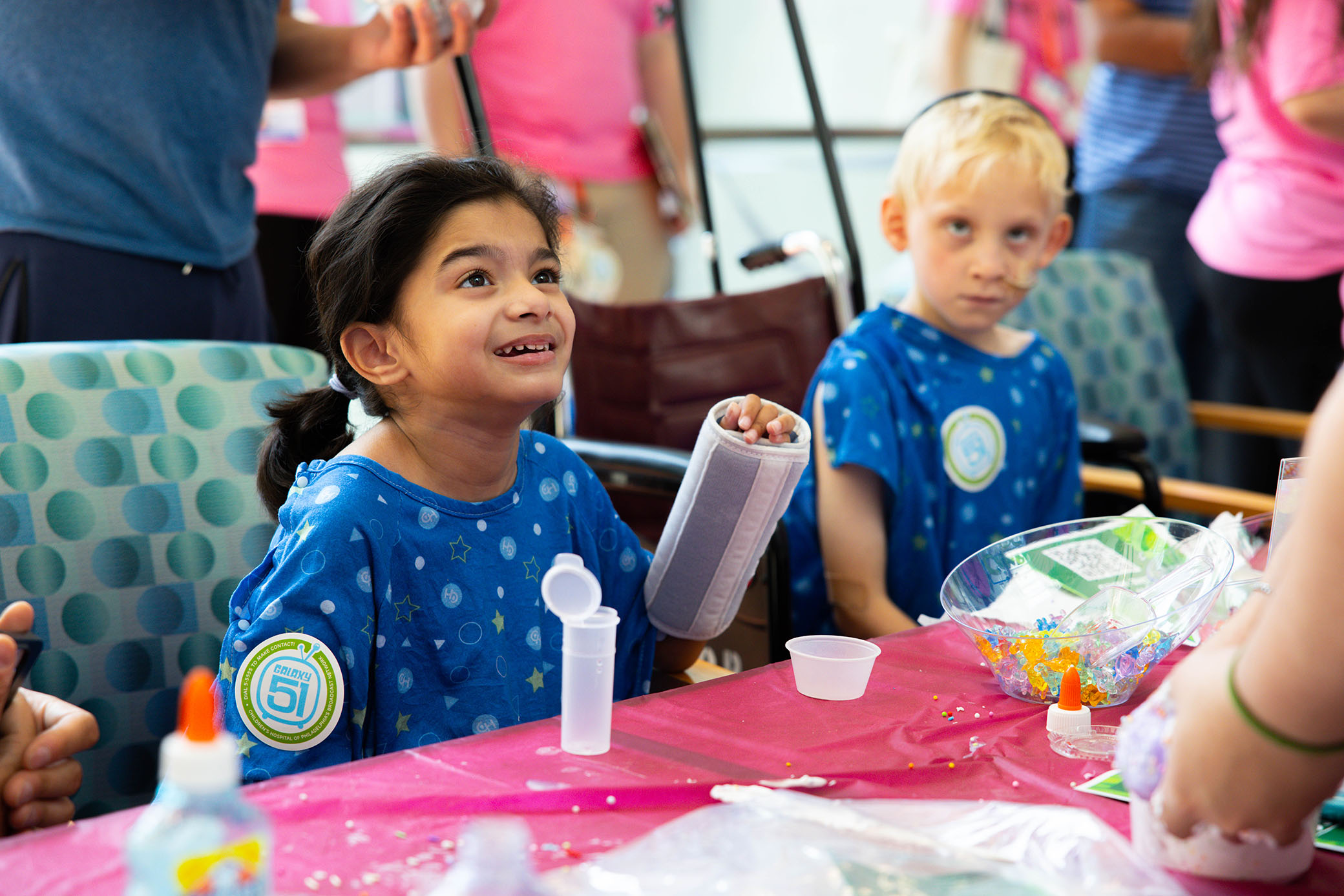 Two children in hospital gowns sit at a craft table covered with colorful supplies, smiling and interacting with others around them in a bright, cheerful room.