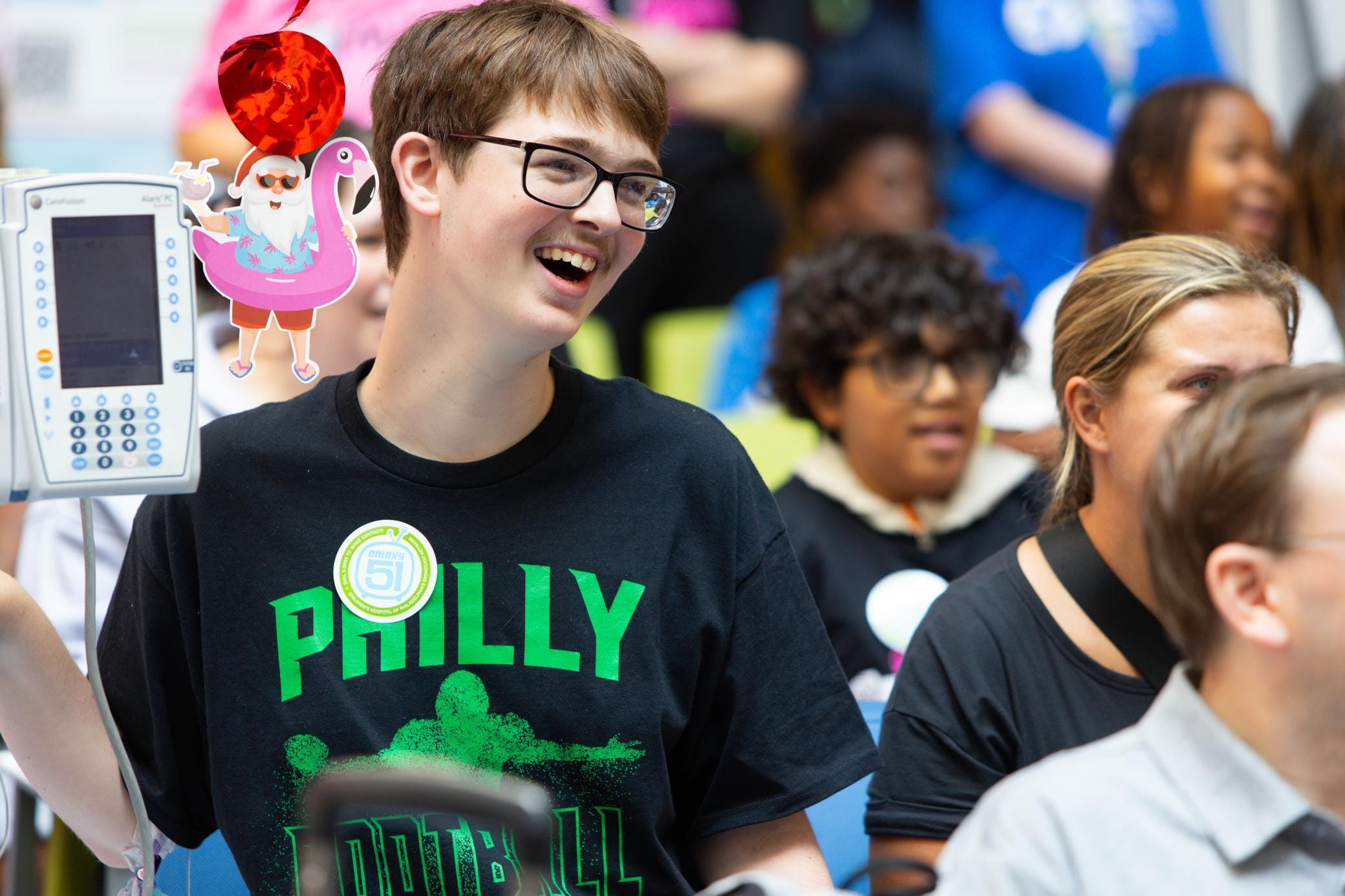 A smiling young person wearing glasses and a Philly Football shirt stands indoors near others, holding a medical device with a flamingo balloon decoration attached. People around them appear engaged and cheerful.