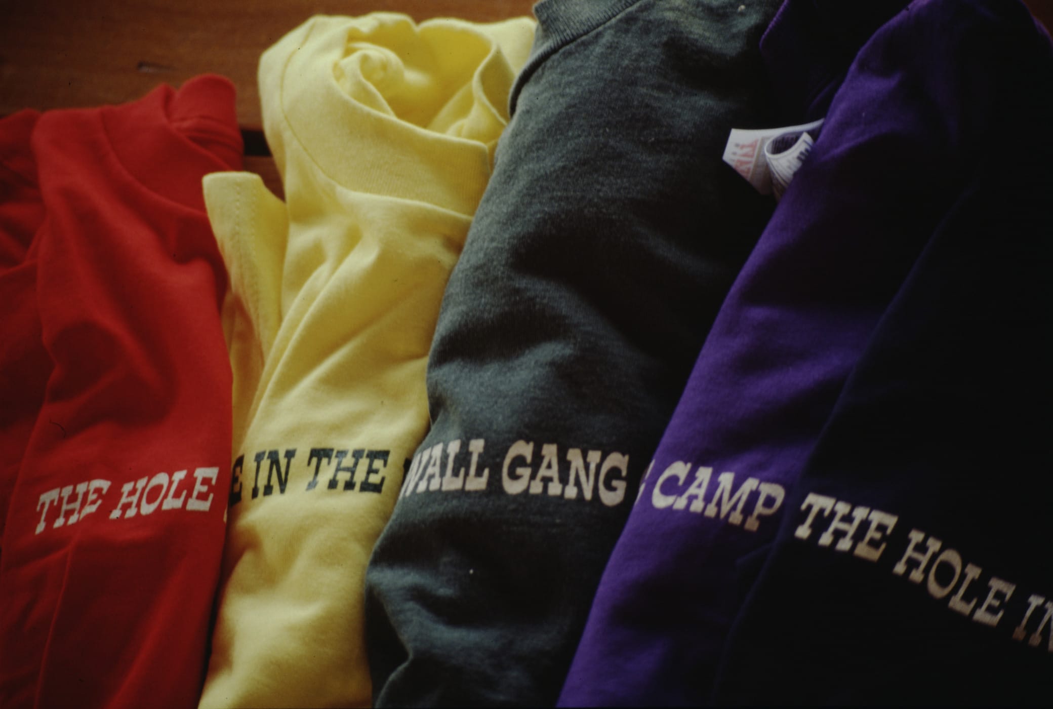Four colorful shirts—red, yellow, dark green, and purple—are laid out side by side. Each shirt features part of the phrase THE HOLE IN THE WALL GANG CAMP printed across the front.