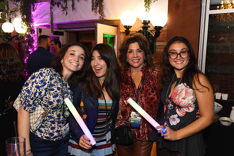 Four women smiling at a party, two holding glowing light sticks. They stand close together with people and festive lights in the background.
