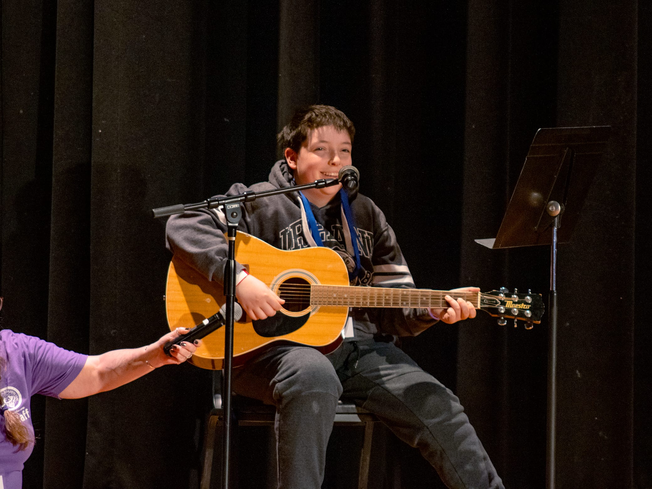 A boy sits on a chair playing an acoustic guitar on stage. He is smiling, wearing a hoodie, and theres a microphone in front of him. A persons arm holds another microphone near the guitar. A music stand is beside him.