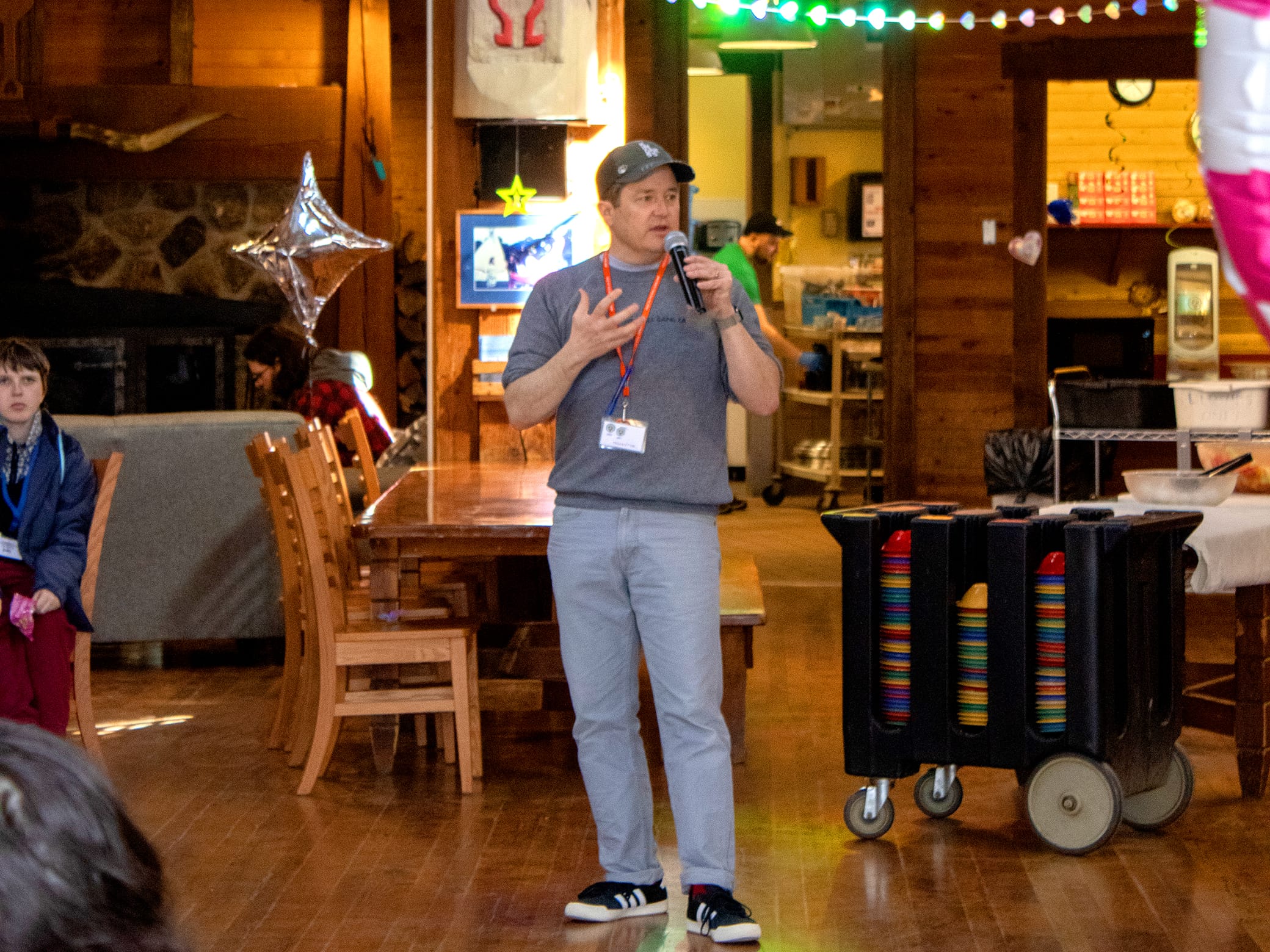 A man speaks with a microphone in a wooden room, wearing a cap and name badge. Chairs, a table, and a disc rack are nearby. People sit and listen. Colorful lights hang overhead.