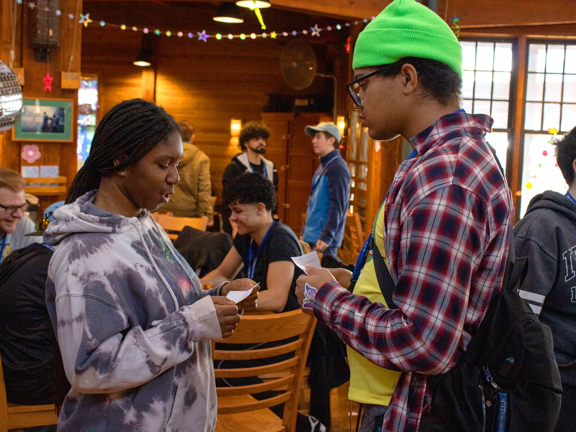 Two people stand facing each other indoors, holding pieces of paper. Other people sit and socialize in the background. The room has wooden walls, string lights, and large windows.