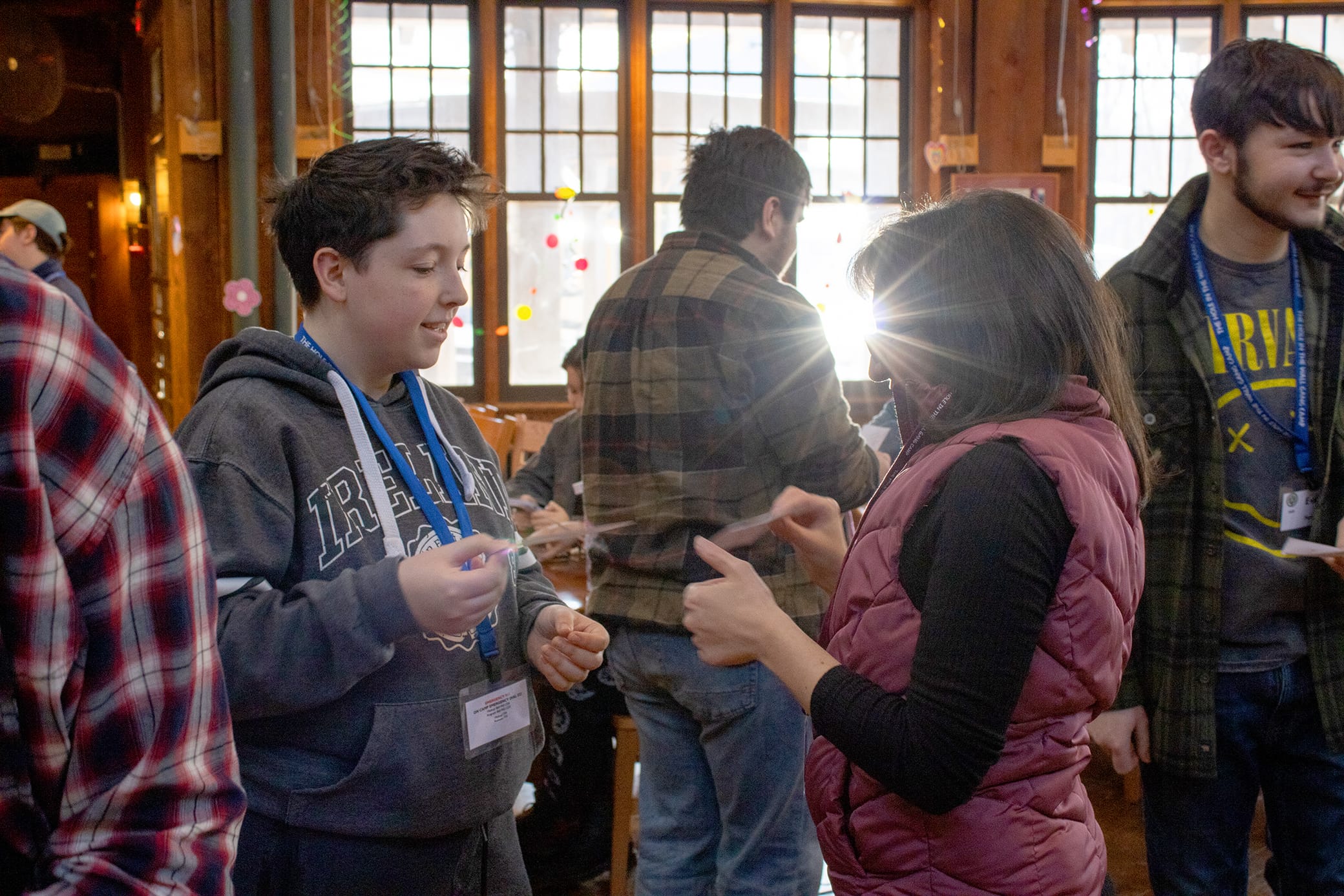 People wearing name tags and winter clothes interact and smile indoors, with sunlight streaming through large windows behind them. Others are gathered and talking in the background of the cozy, wood-paneled room.